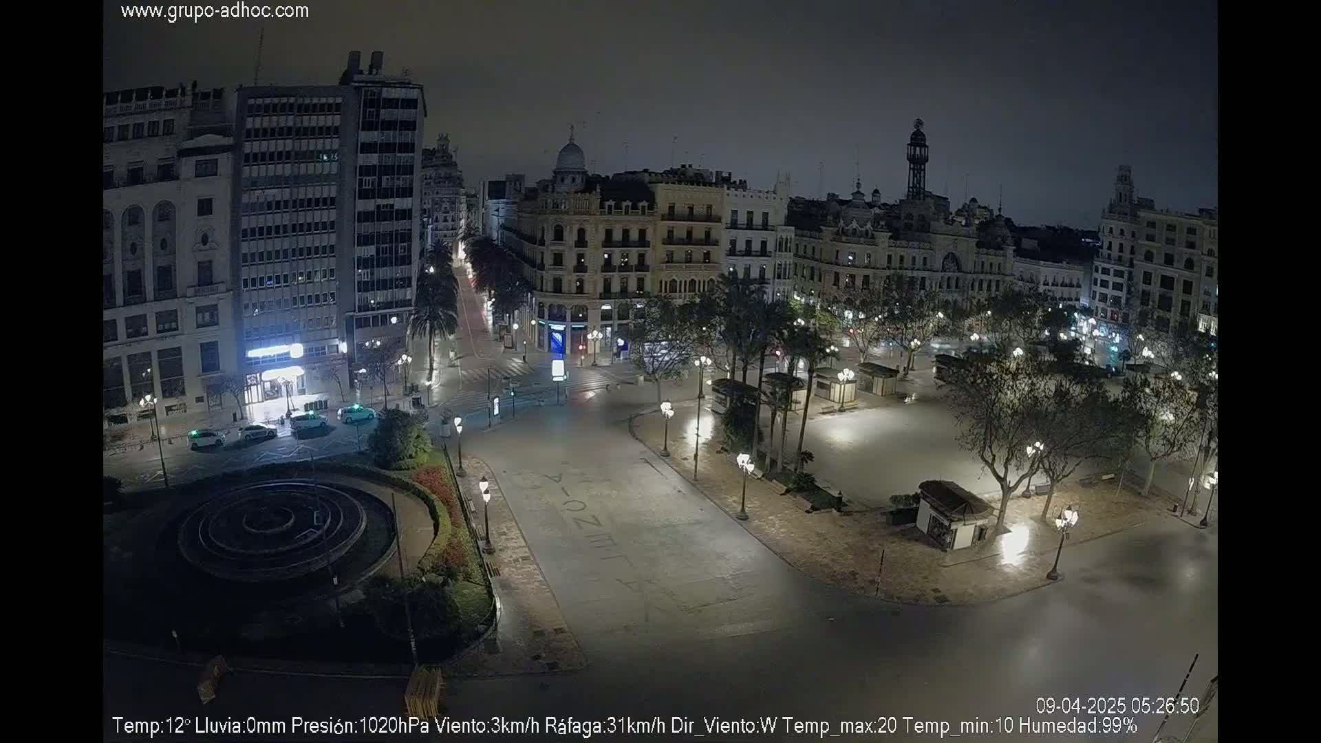 A mostly empty, brightly lit city square at night, surrounded by tall buildings under a clear, dark sky.