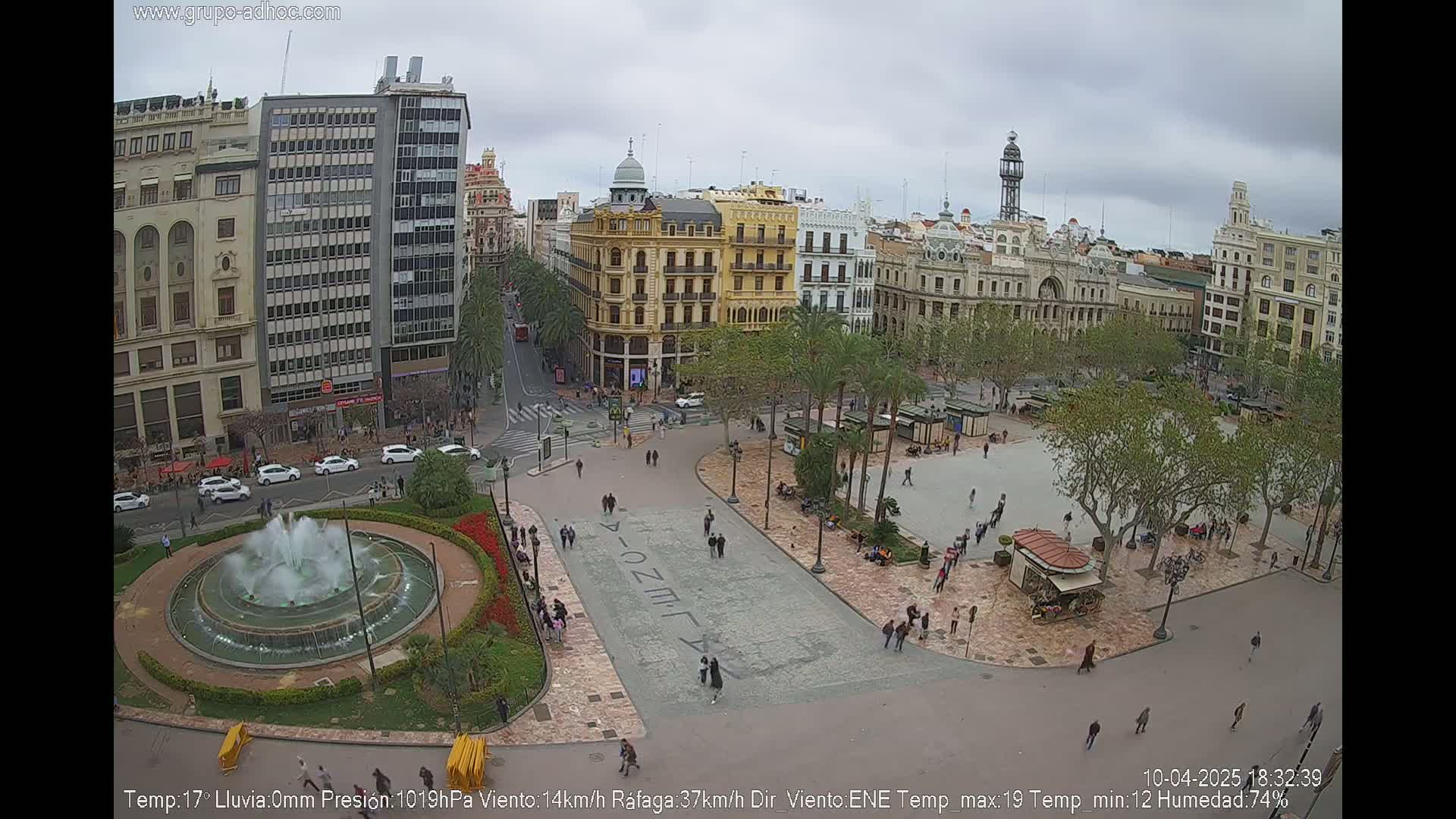 An elevated view of a city square with a fountain, palm trees, and several buildings under an overcast sky.