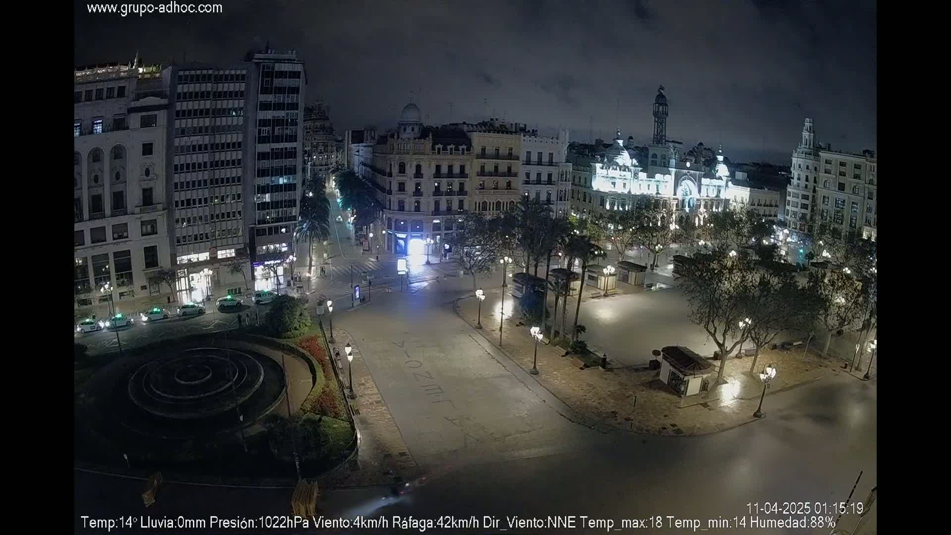 A nighttime view of a city square with buildings surrounding it, illuminated with streetlights under a cloudy sky.