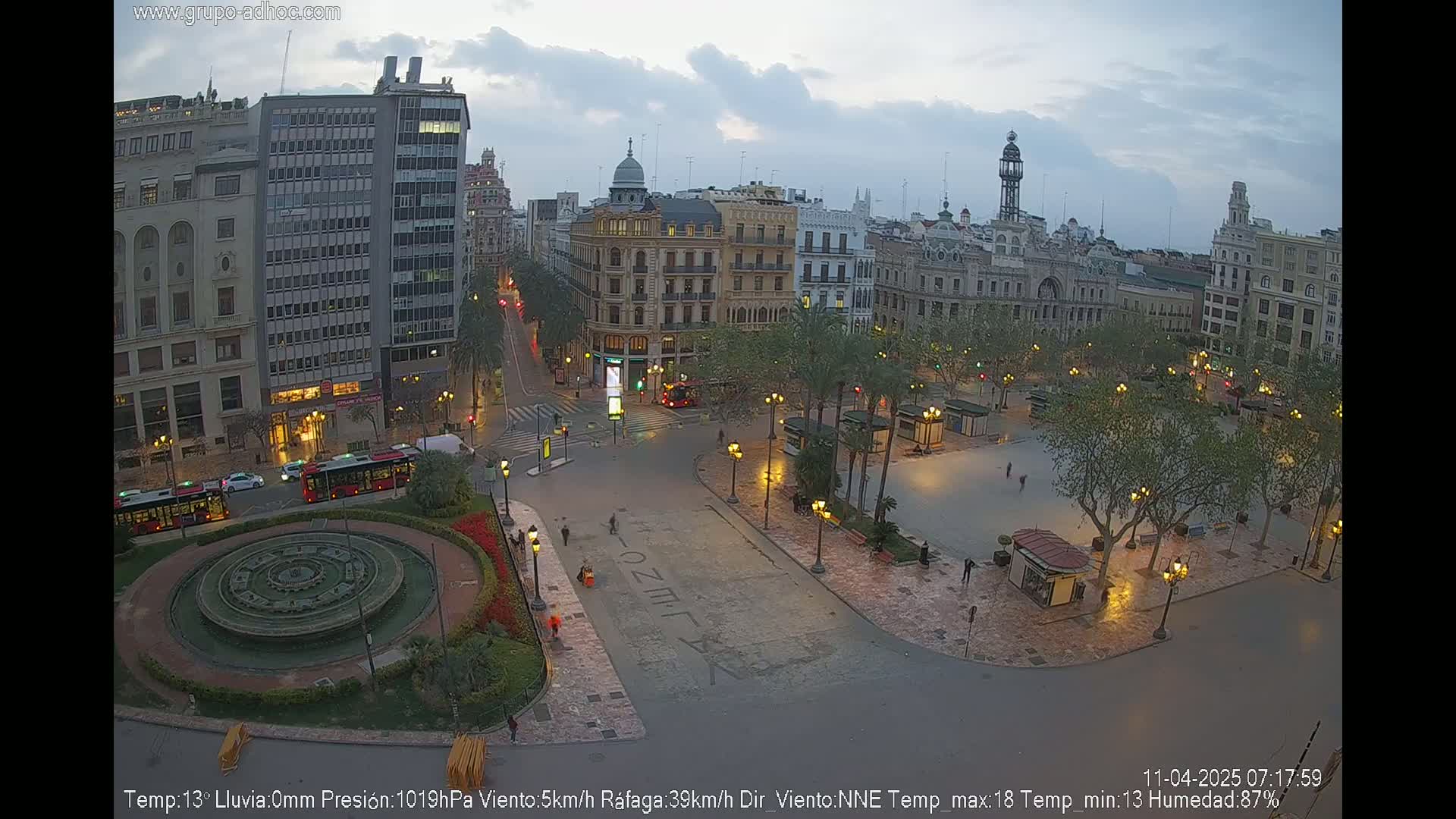 A city square at dusk, under a cloudy sky, features a fountain, palm trees, and buildings illuminated by streetlights.