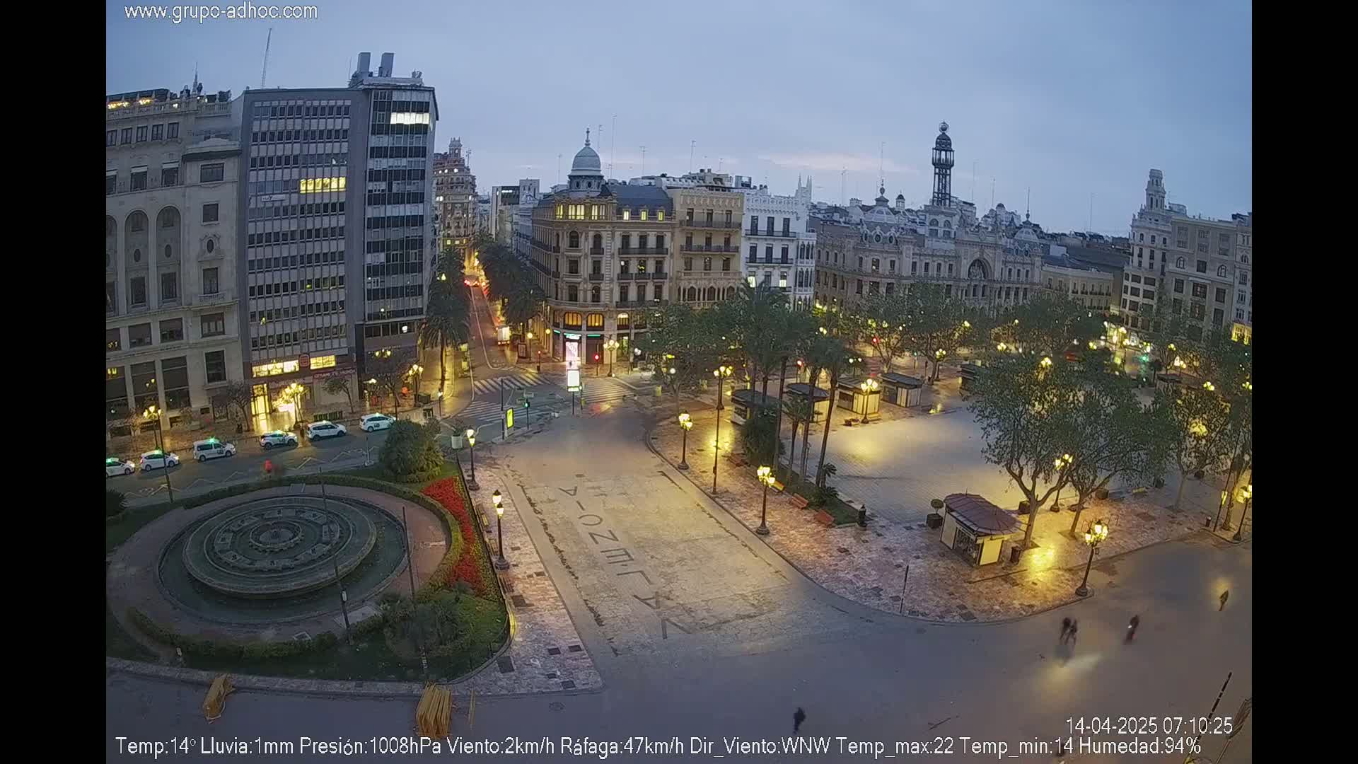 A city square at night, illuminated by streetlights, with a fountain in the foreground and light rain falling.