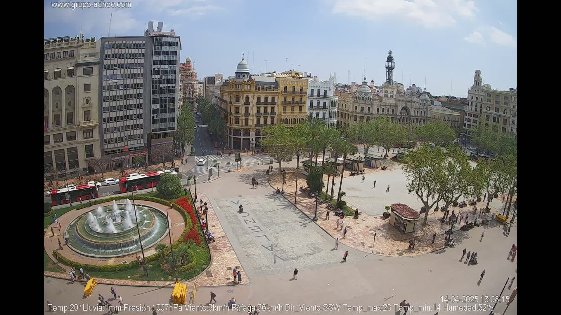 A city square with a fountain, surrounded by buildings and palm trees, on a partly sunny day with light rain.