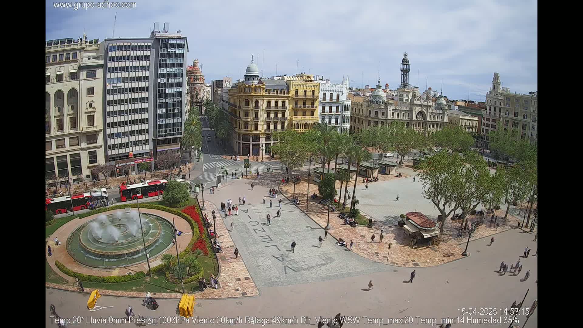A city square with a fountain, surrounded by buildings and palm trees under a partly cloudy sky.