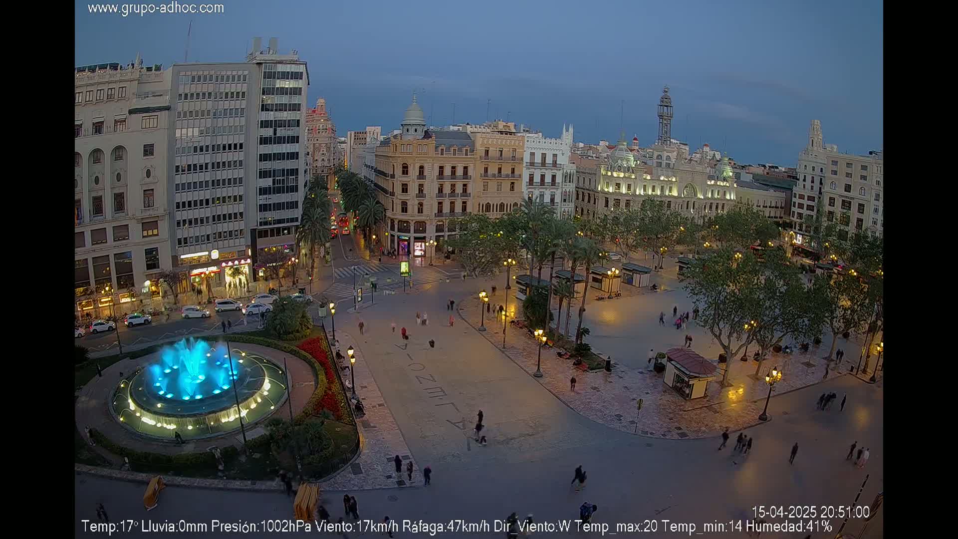 A nighttime aerial view of a city square with a lit fountain, surrounded by buildings and palm trees under a twilight sky.