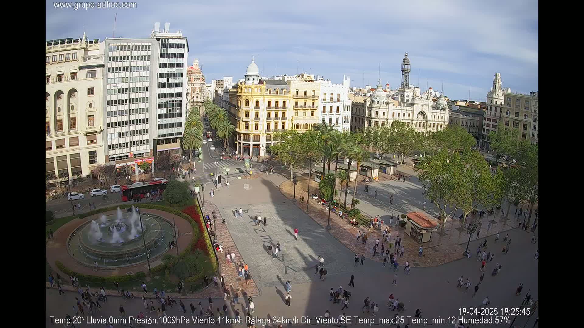 A sunny city square, populated with pedestrians and palm trees, features a central fountain and surrounding buildings.