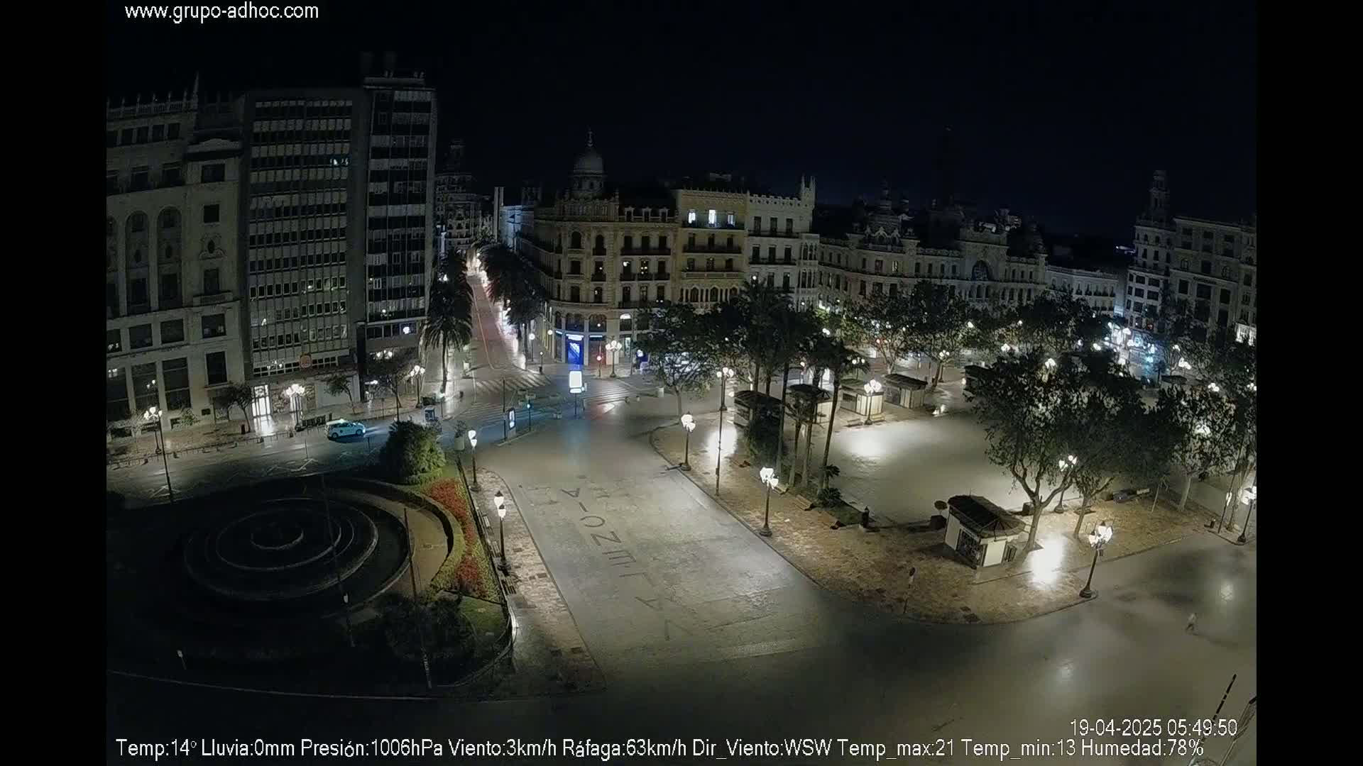 A nighttime aerial view of a city square with buildings illuminated, featuring a circular fountain and sparsely populated streets under clear skies.