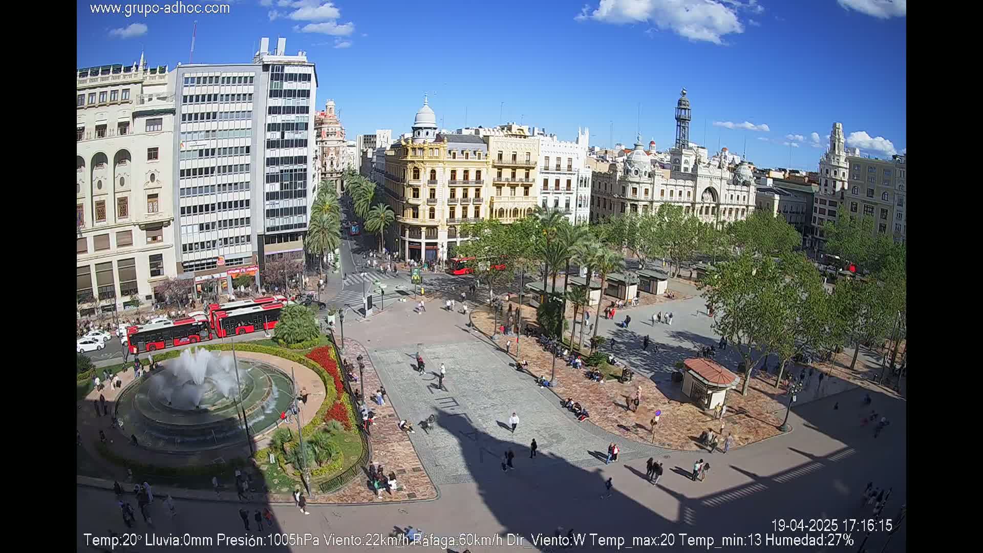 A sunny city square with a fountain, palm trees, and people relaxing on a warm day.
