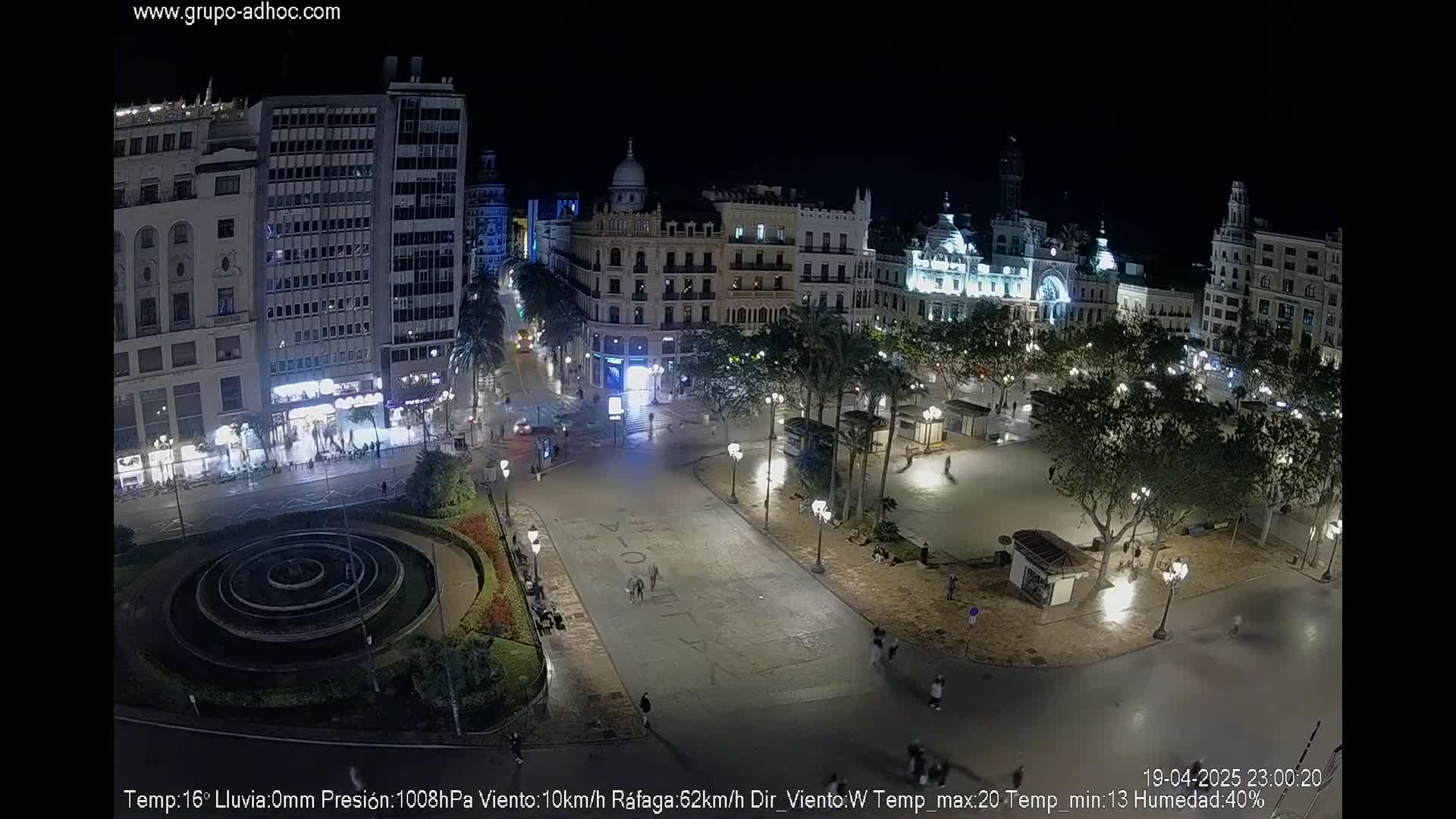 A nighttime aerial view of a city square with surrounding buildings, palm trees, and a circular fountain, under clear skies.