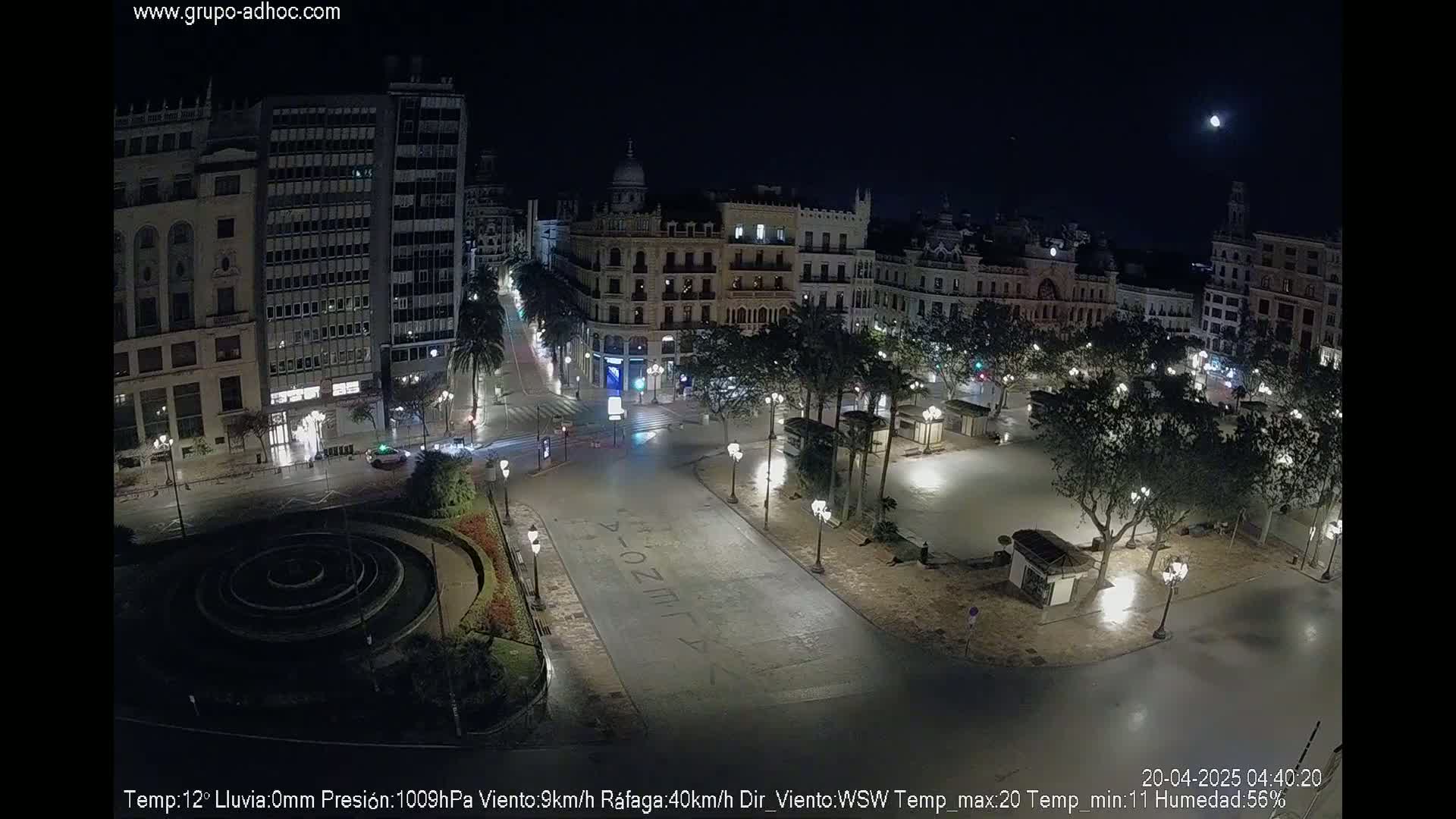 A mostly empty, nighttime plaza with a circular fountain in the center, surrounded by tall buildings under a clear night sky with a visible moon.
