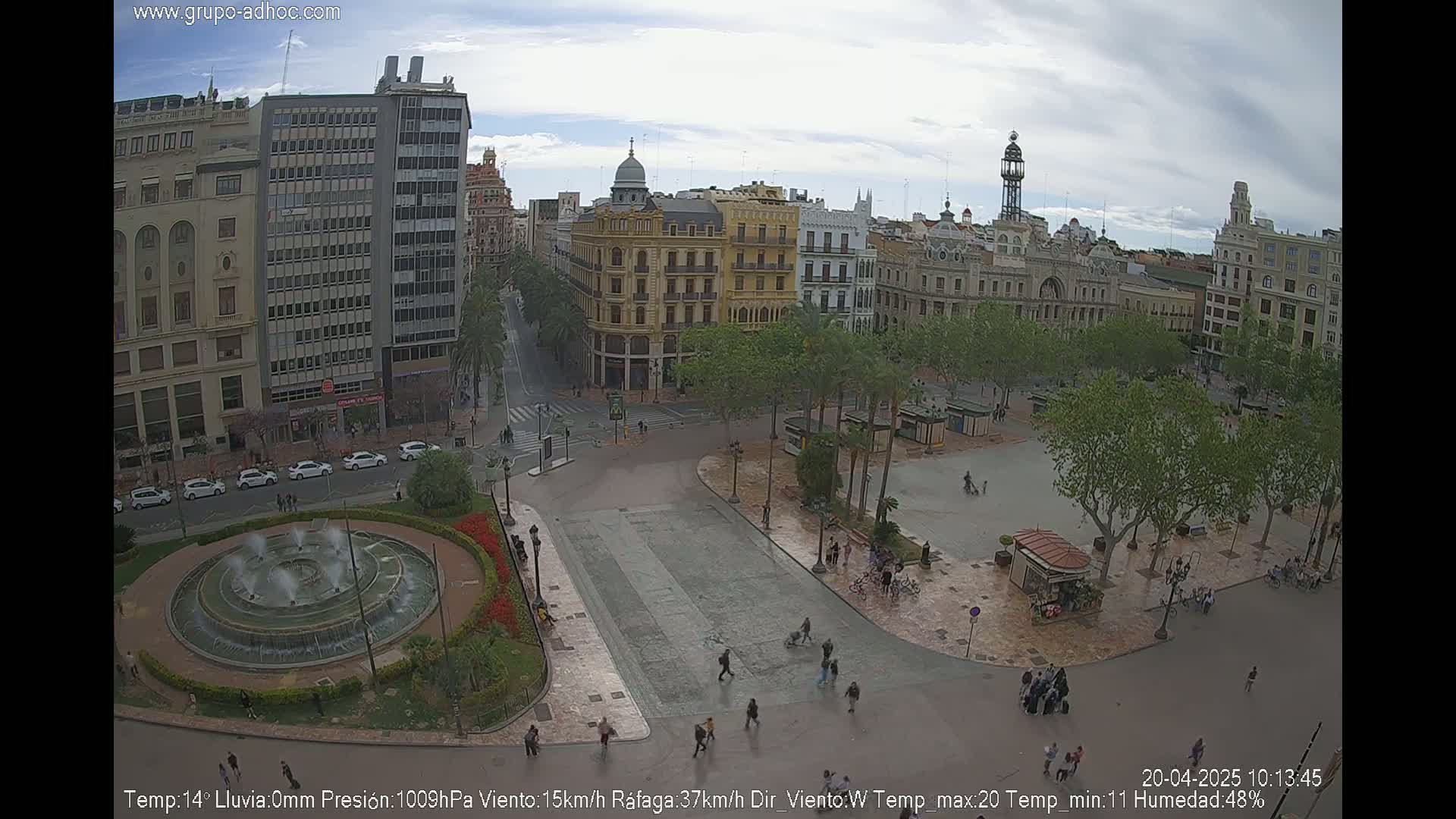 A city square with a fountain, surrounded by buildings and trees under a partly cloudy sky.