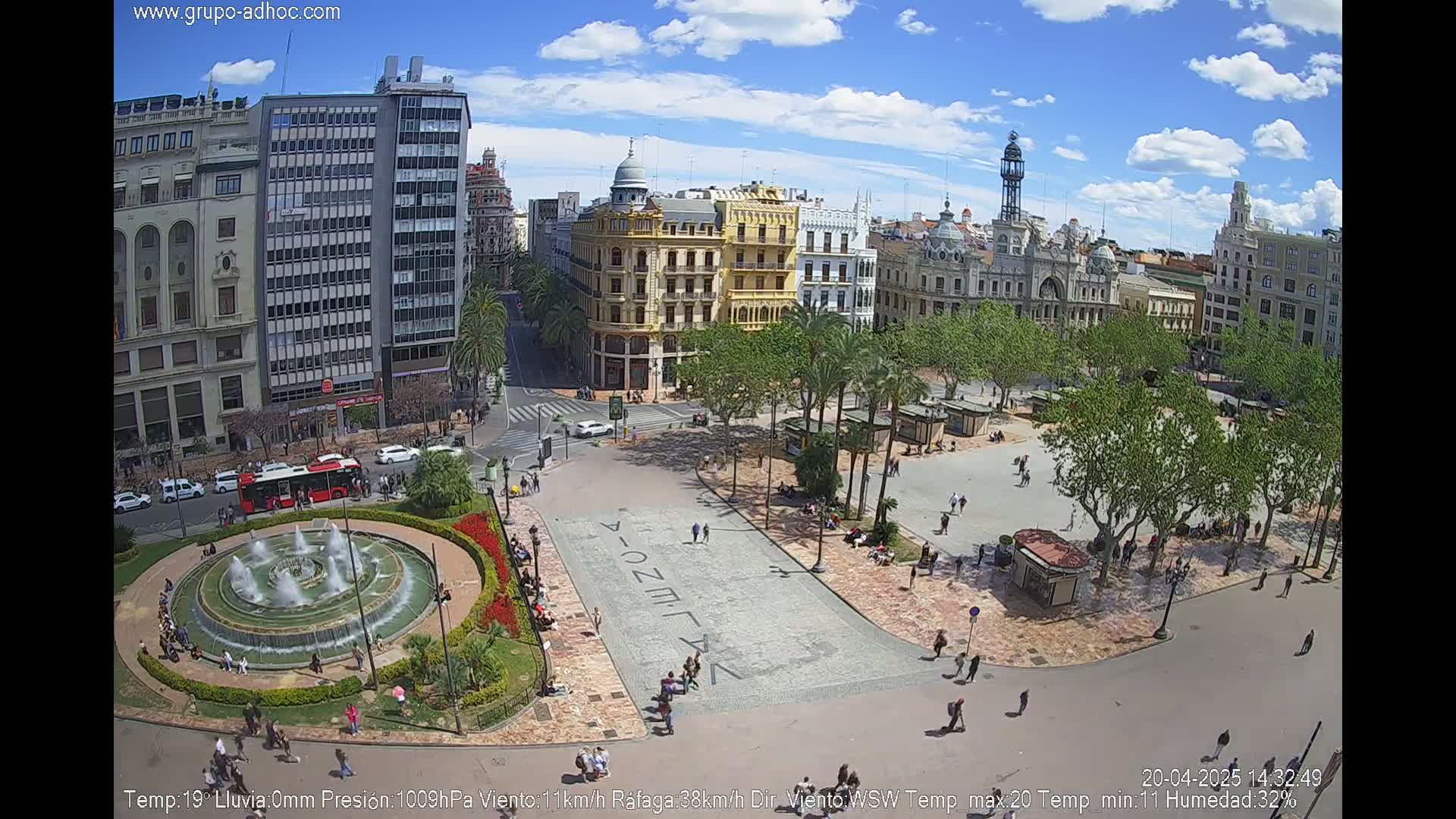 A city square with a fountain, surrounded by buildings and palm trees, under a partly sunny sky.