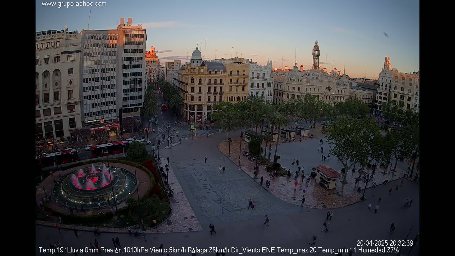 A city square at dusk, under a clear sky, features a central fountain, surrounded by people walking near buildings and palm trees.