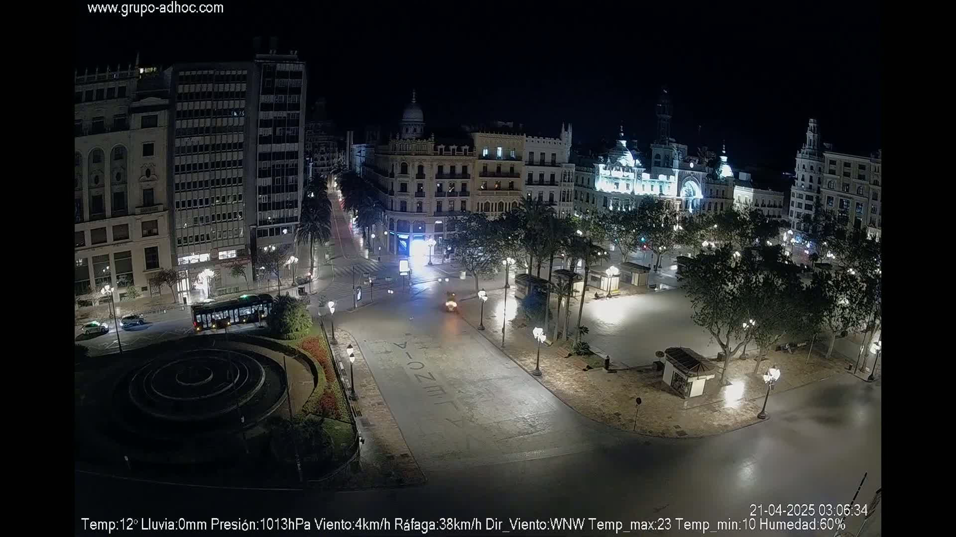 A nighttime, high-angle view of a mostly empty city square with buildings illuminated, and a few vehicles on the streets, under partly cloudy skies.