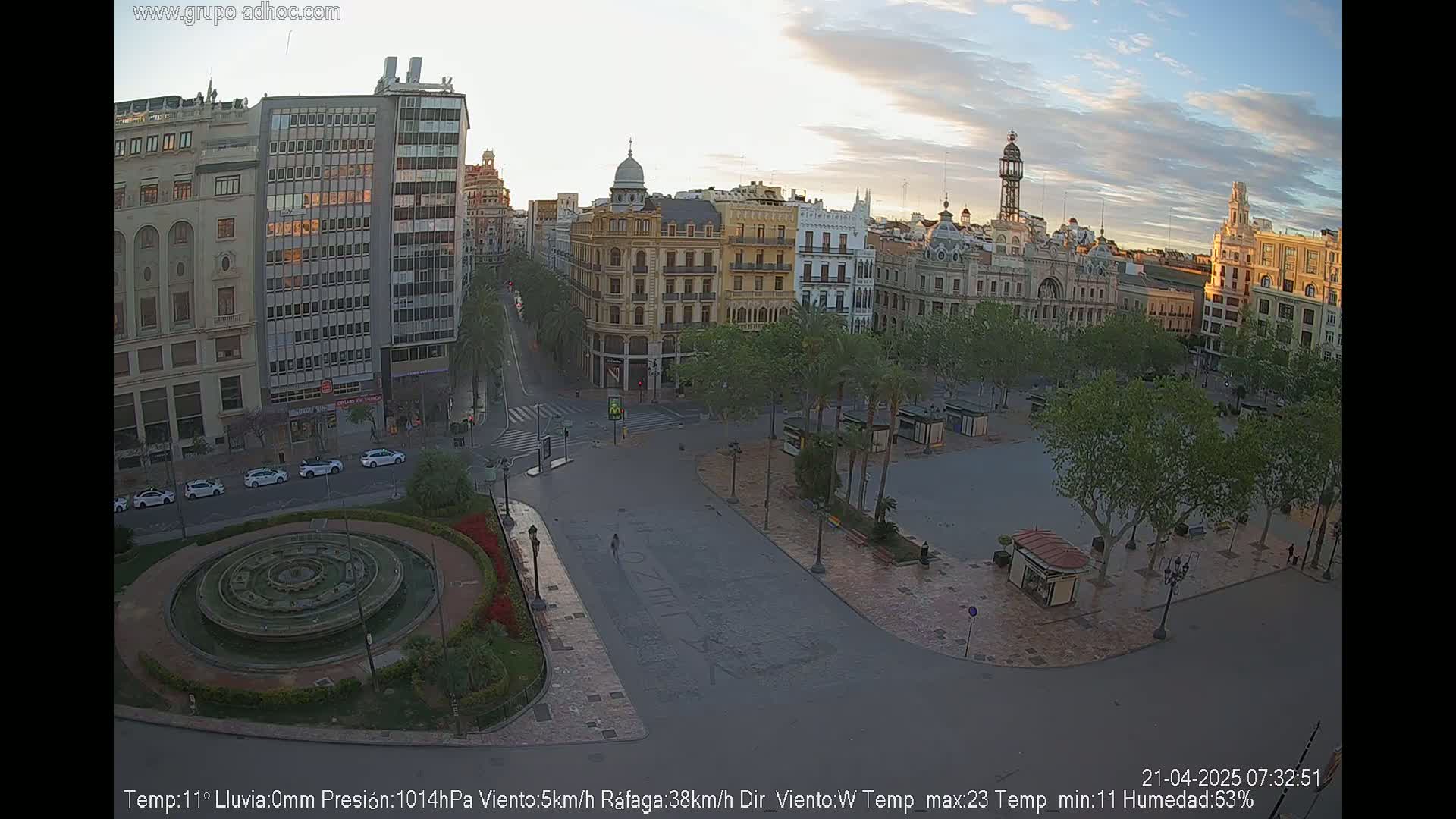 A high-angle, long shot shows a city square at sunrise, with several buildings surrounding it, a central fountain, and a few cars parked on the streets.