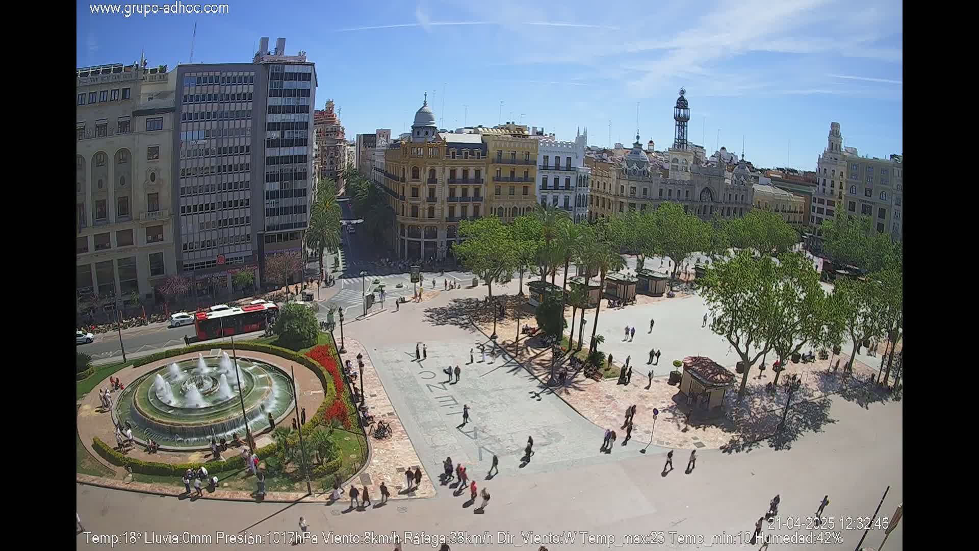 A city square with a fountain, surrounded by buildings and trees under a sunny sky.