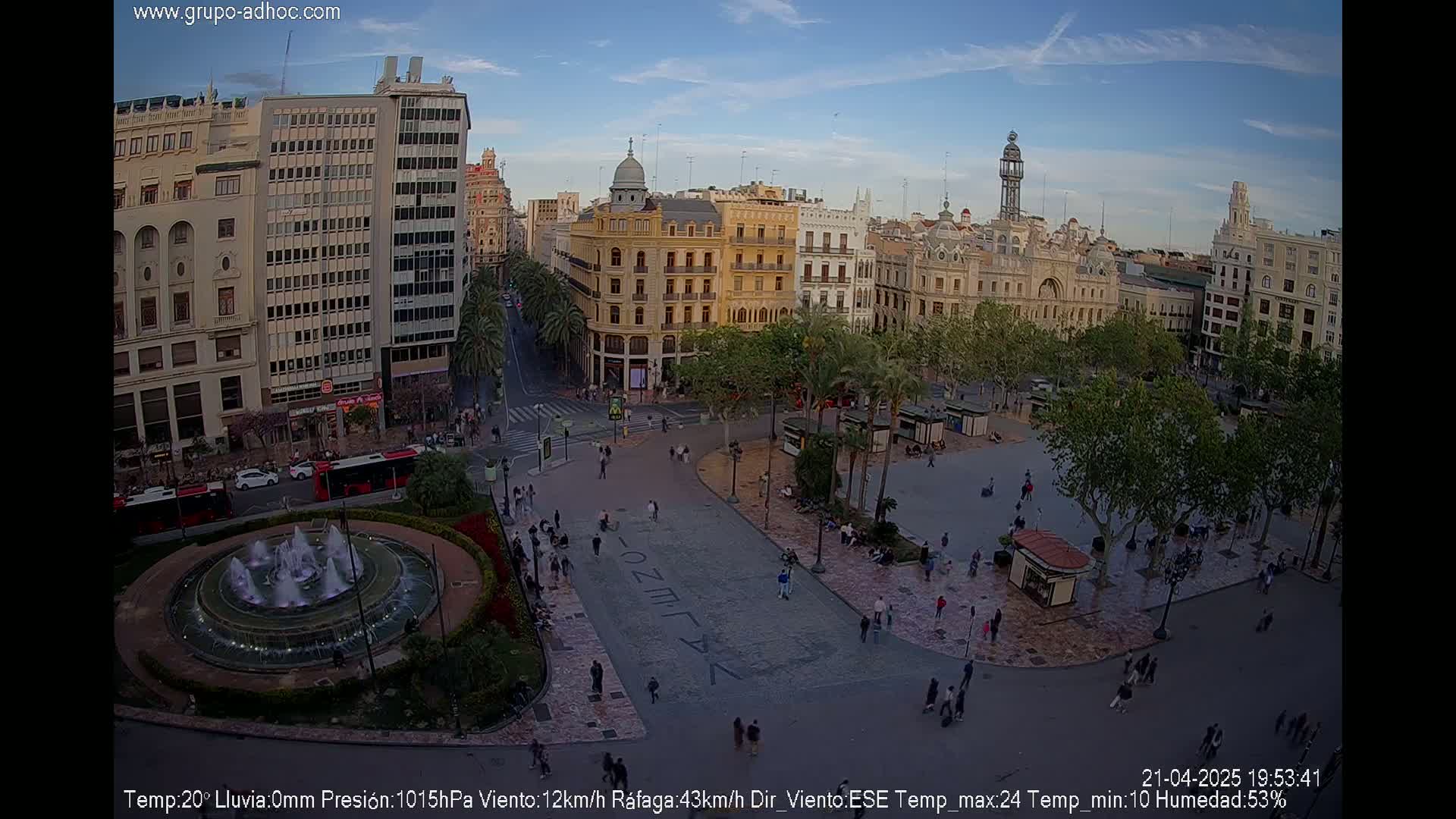 A city square with a fountain, surrounded by buildings under a partly cloudy sky.