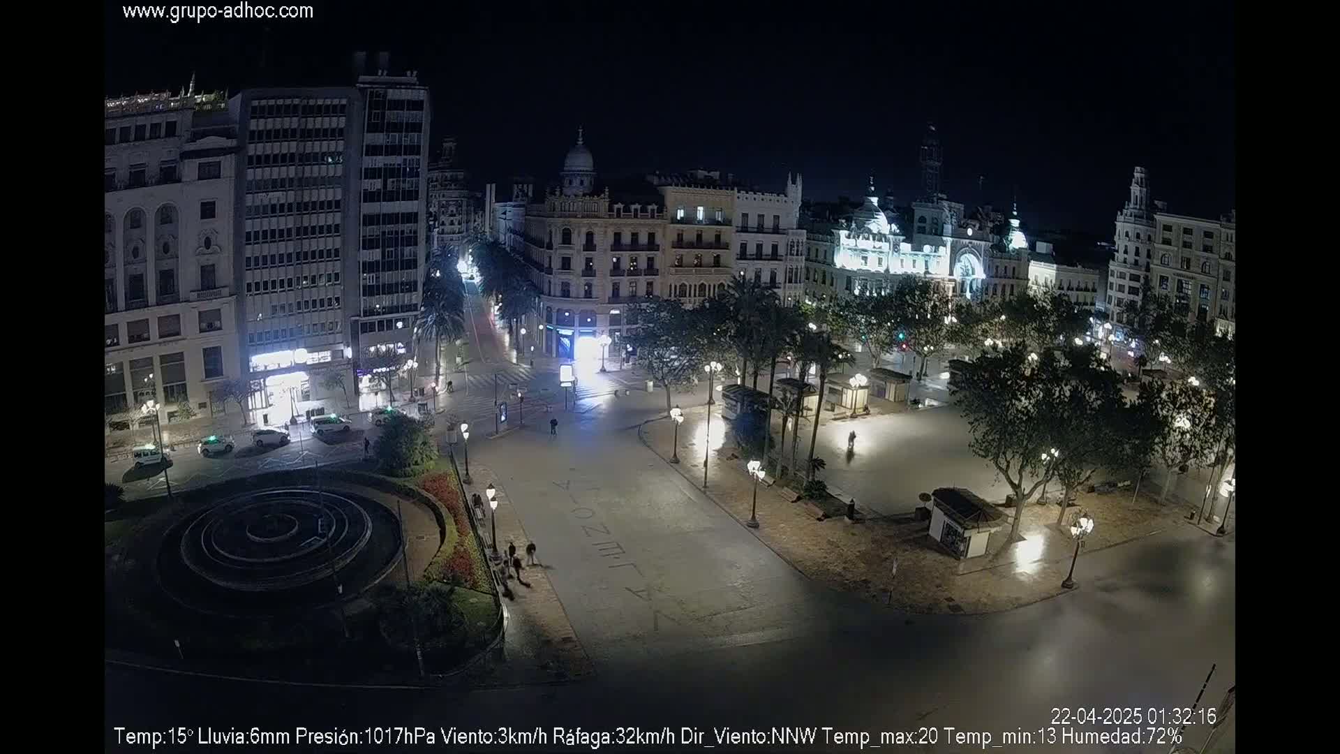 A nighttime, aerial view of a city square with several buildings, trees, and a circular fountain, under rainy conditions.