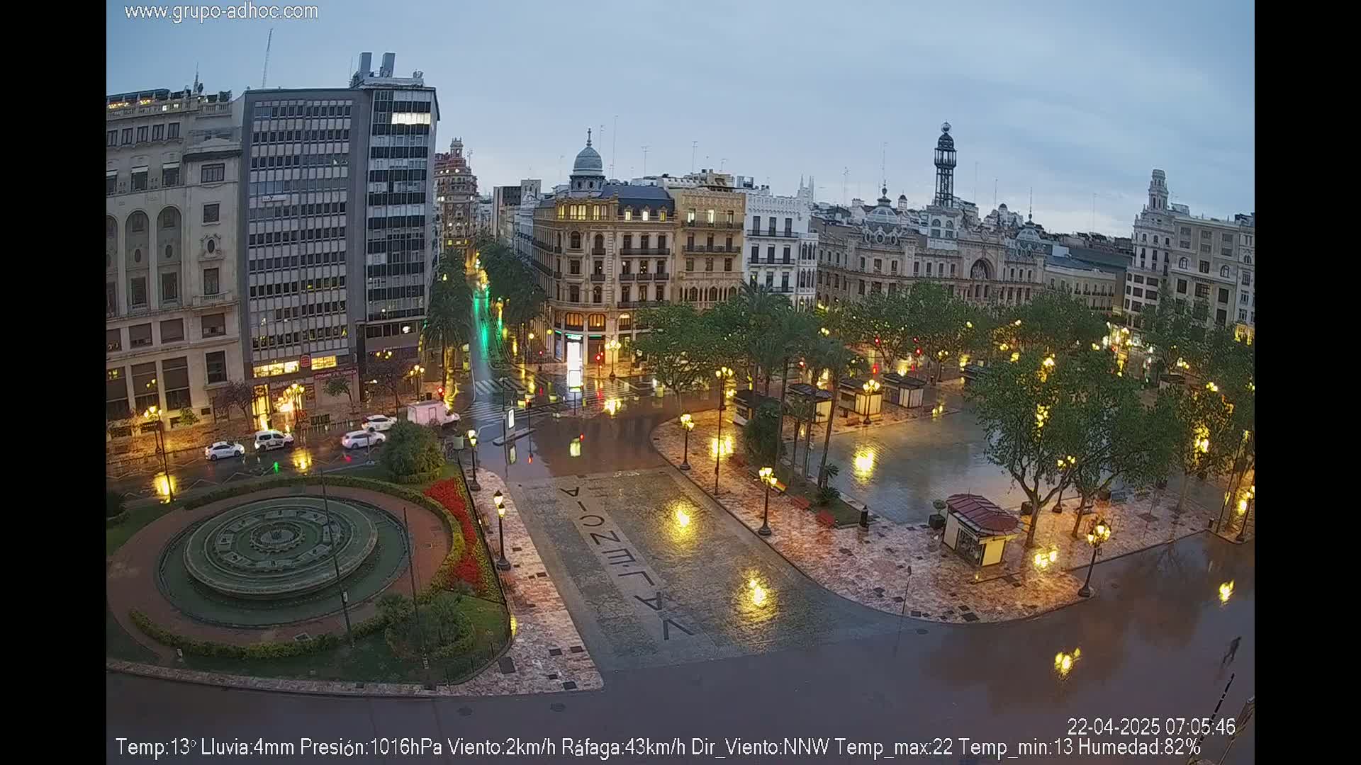 A rain-soaked city square at night, illuminated by streetlights, features a circular fountain and surrounding buildings.