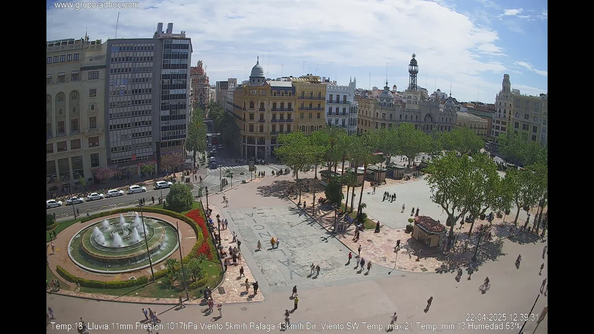 A city square with a fountain, surrounded by buildings and trees under a partly cloudy sky.