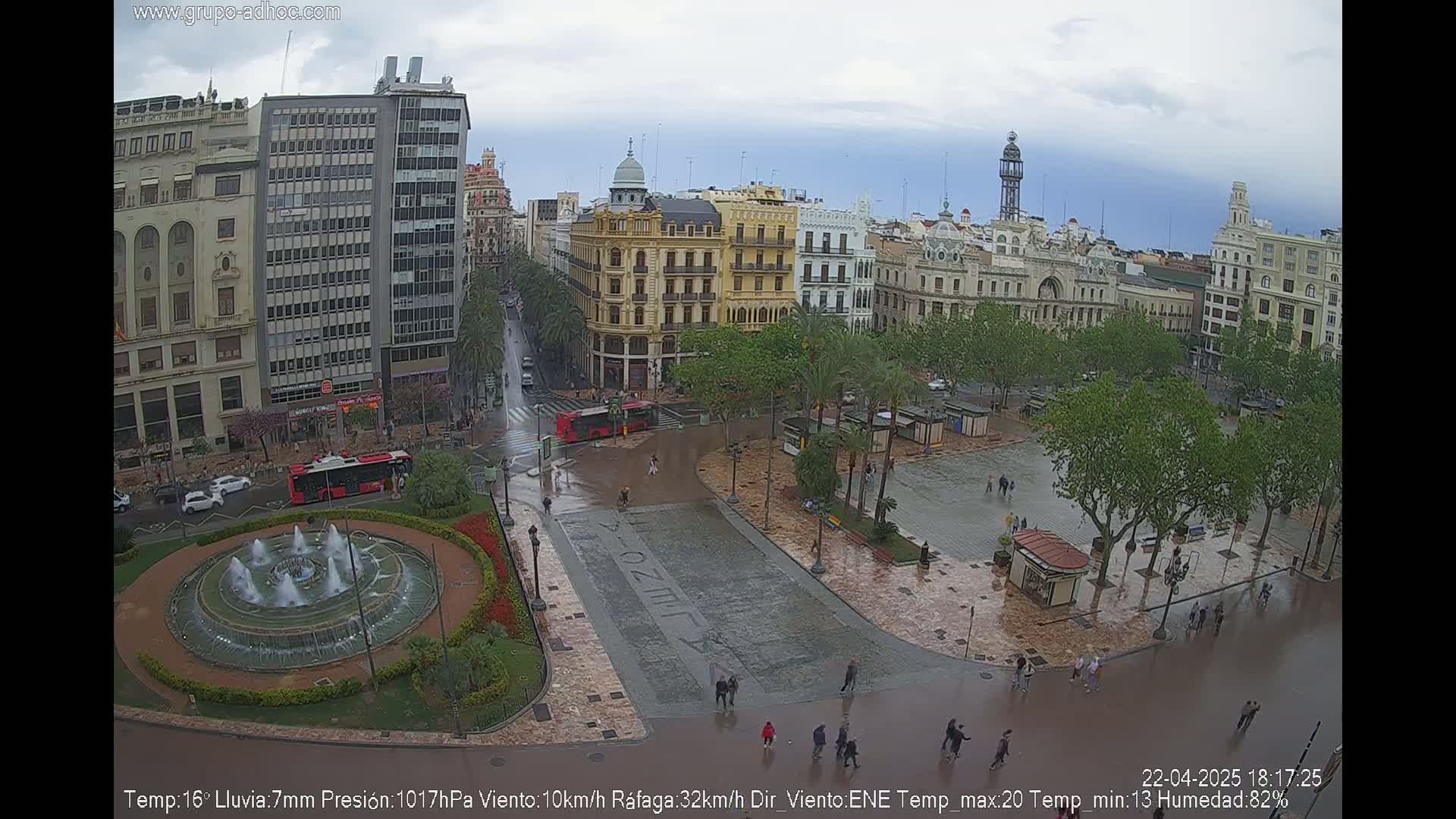 A city square, wet from rain, is surrounded by buildings and trees, with a fountain in the center.