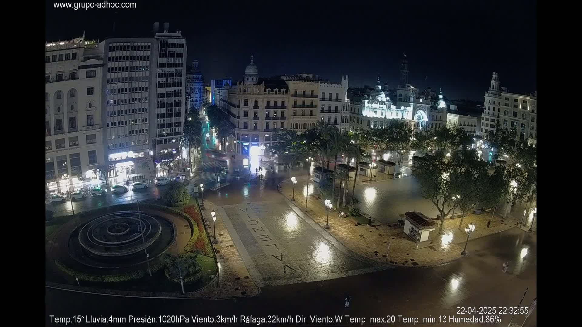 A rain-slicked city square at night, surrounded by buildings, with a circular fountain in the center.