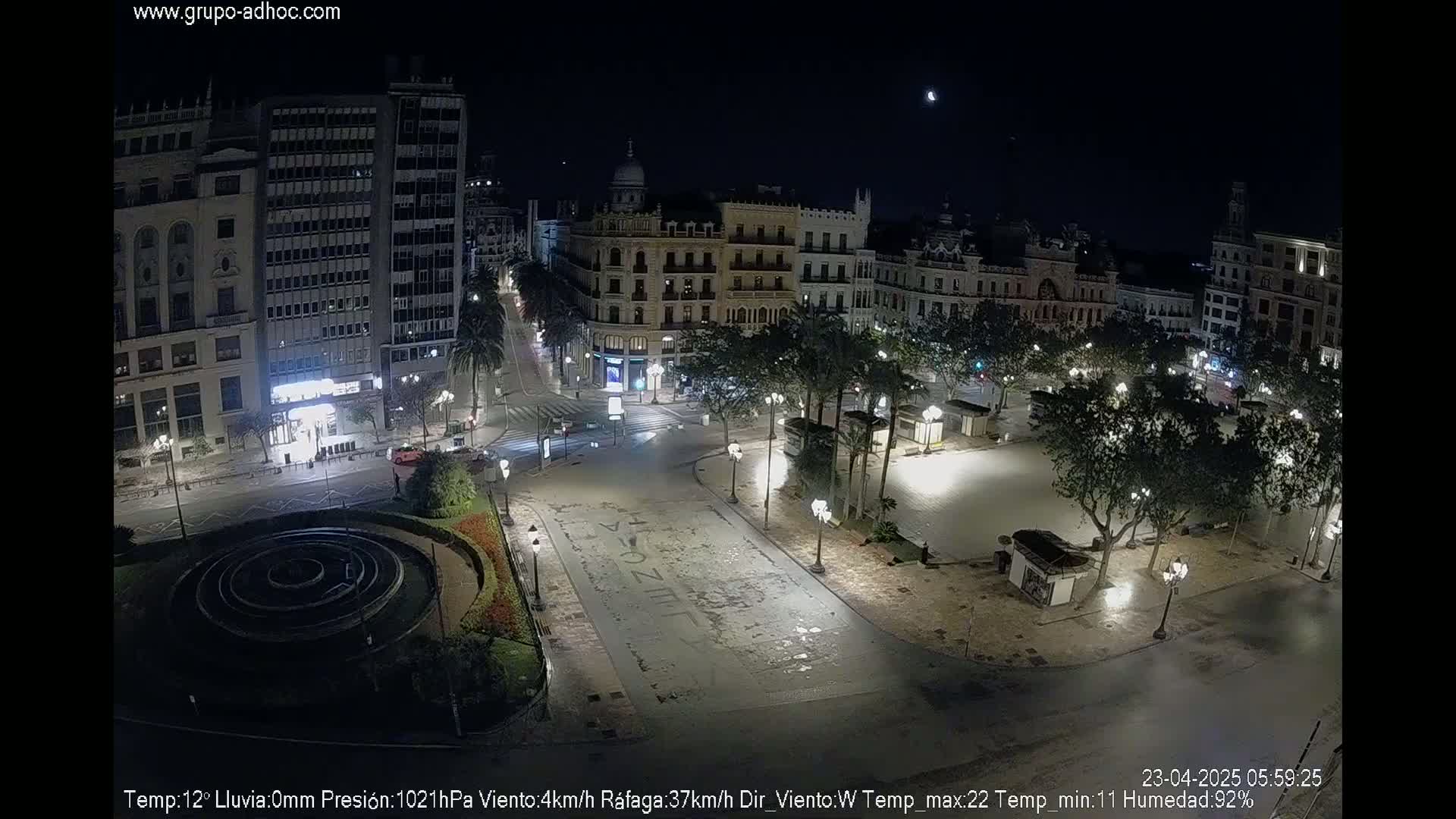 A nighttime view of a city square, illuminated by streetlights, with buildings surrounding it under a clear sky and a visible moon.