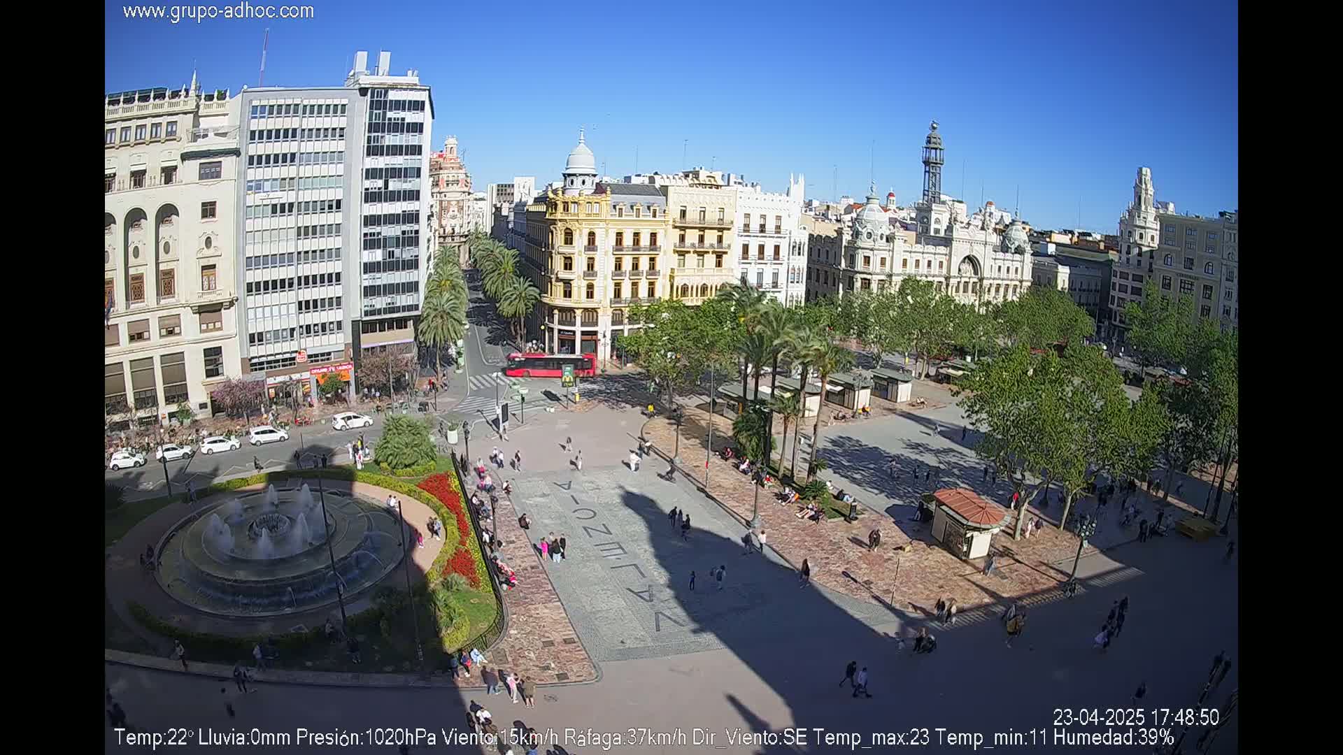 A sunny city square with a fountain, surrounded by buildings, palm trees, and people enjoying the day.
