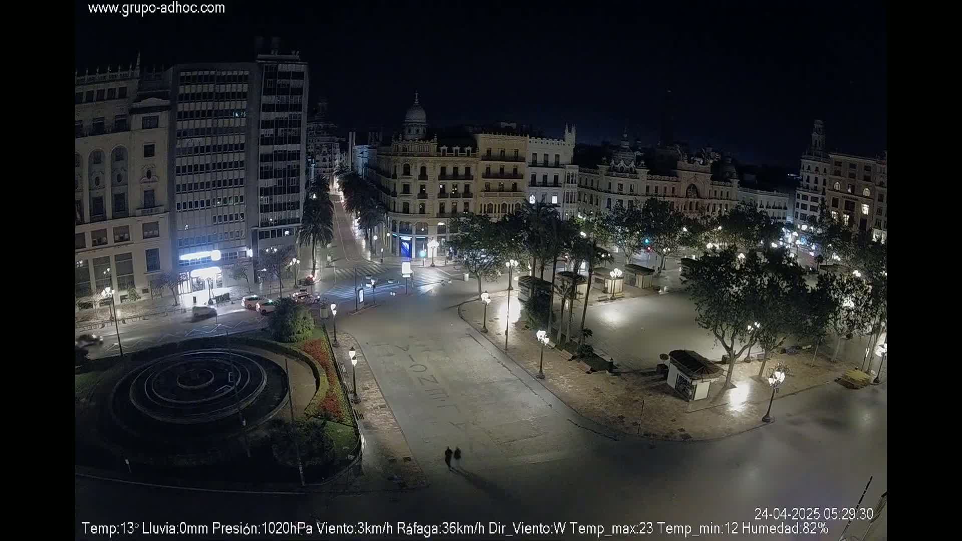 A mostly empty city square at night, illuminated by streetlights, with several buildings visible in the background under a clear, dark sky.