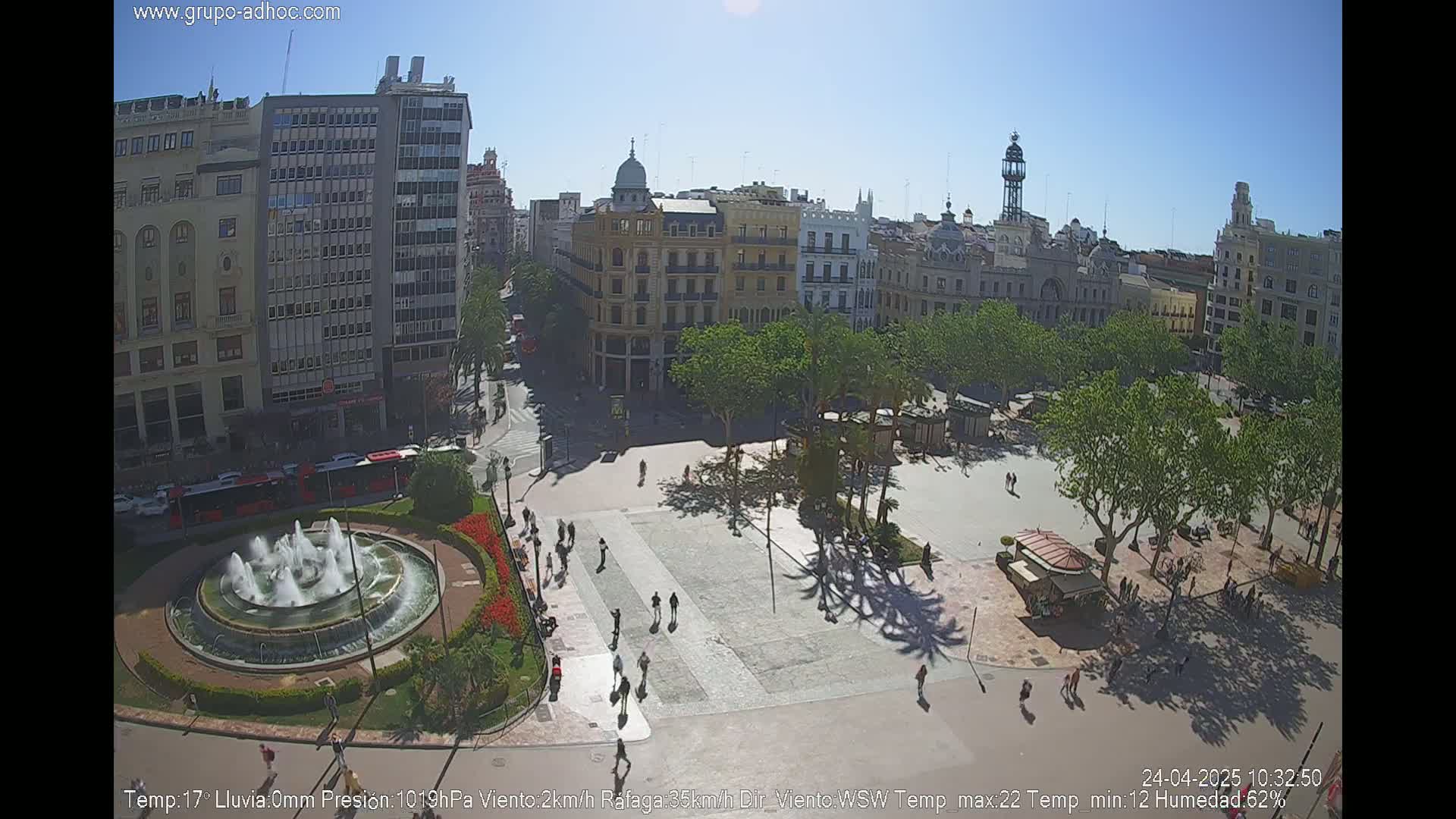 A sunny city square with a fountain, surrounded by buildings and trees, shows people walking on a clear day.