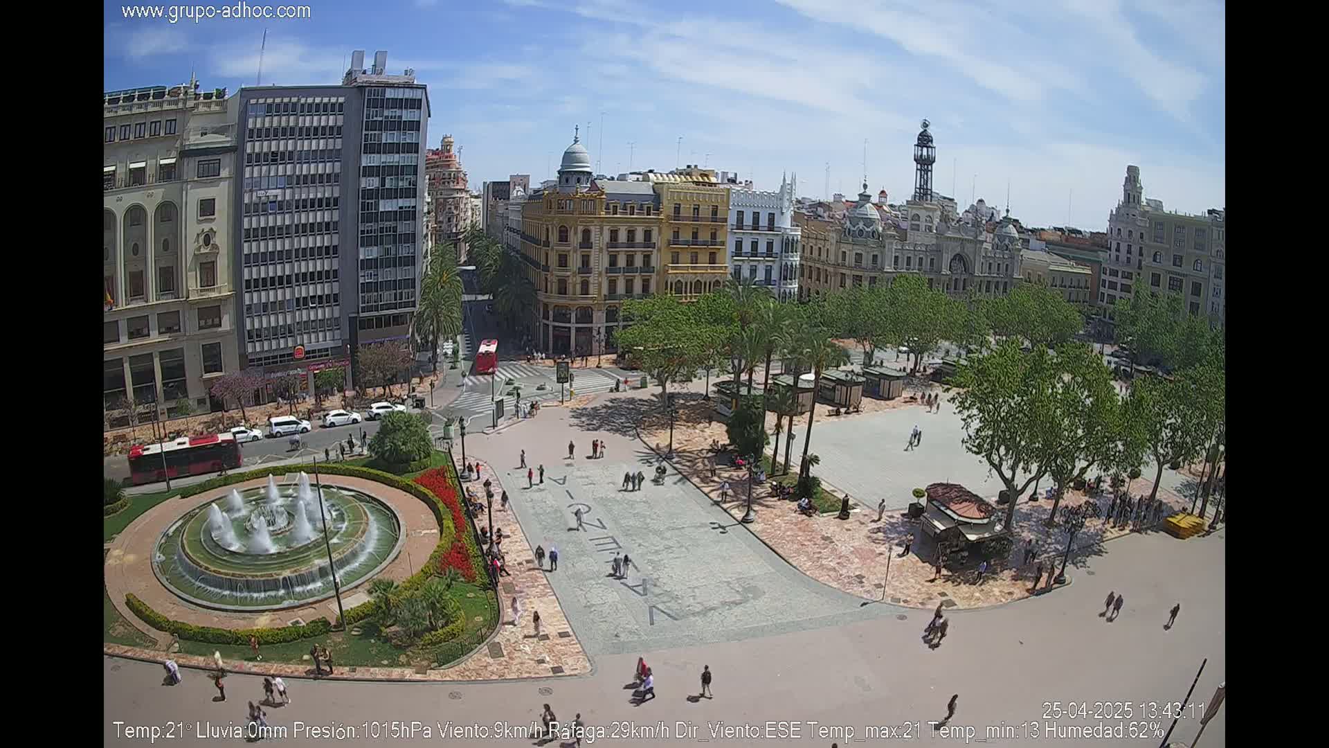 A sunny city square with a fountain, trees, and people walking, surrounded by buildings.