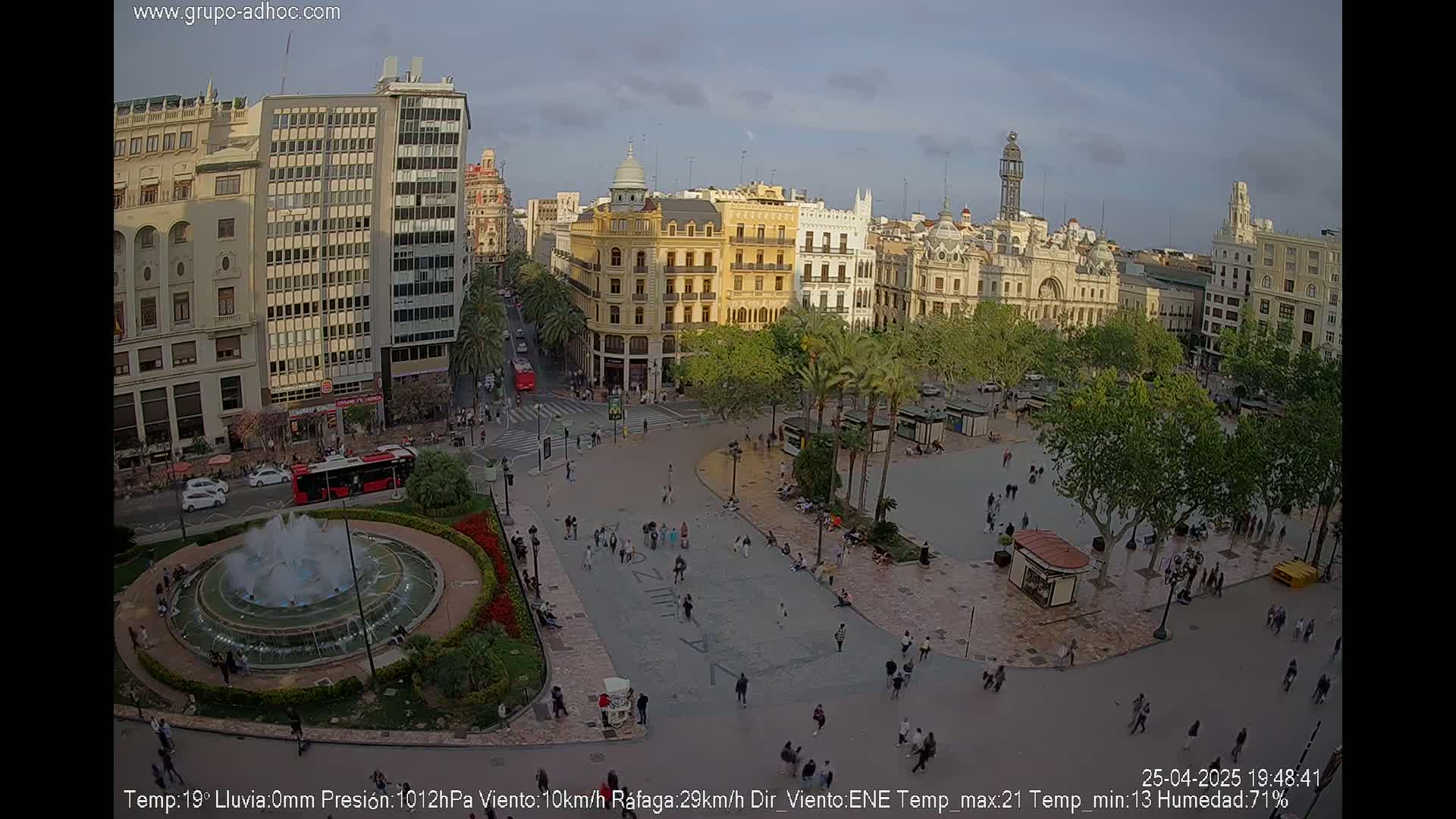 A city square with a fountain and numerous people milling about, under a partly cloudy sky.