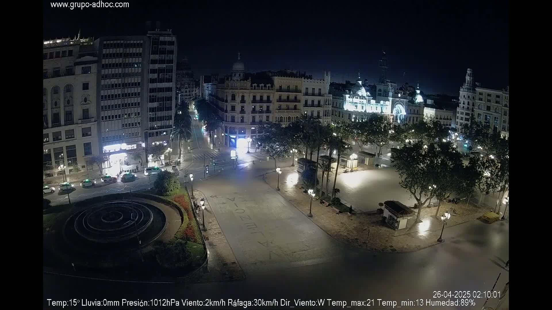 A nighttime view of a city square, illuminated by streetlights, with several buildings surrounding it and a circular fountain in the foreground.