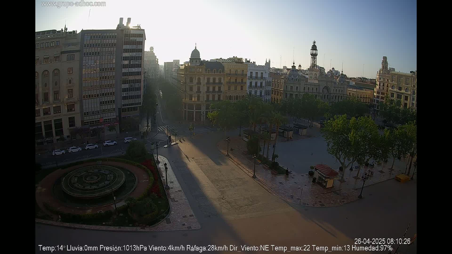 A high-angle, long shot of a city square with a fountain, buildings, and trees on a sunny, clear morning.