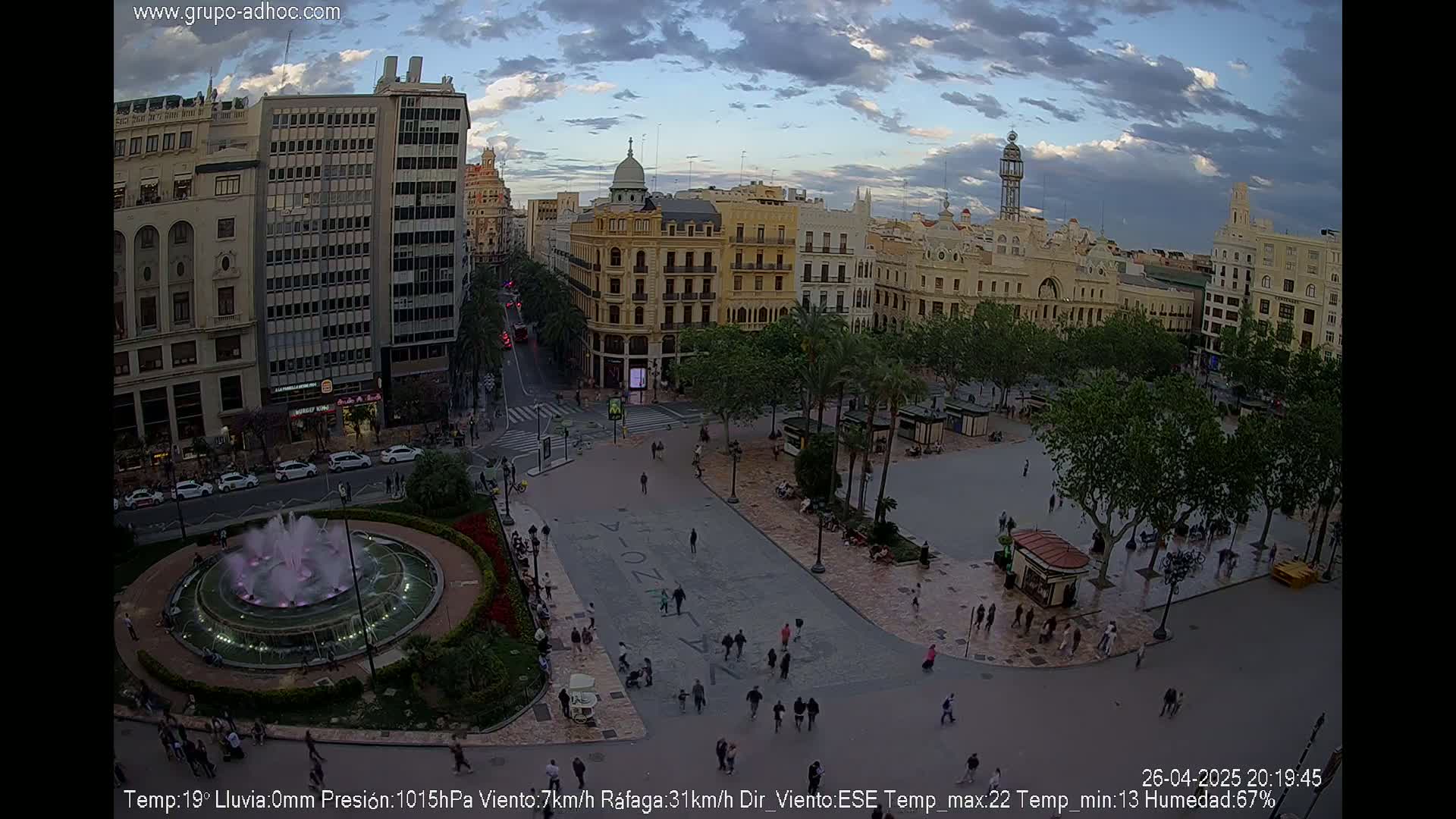 A city square with a fountain, surrounded by buildings under a partly cloudy evening sky.