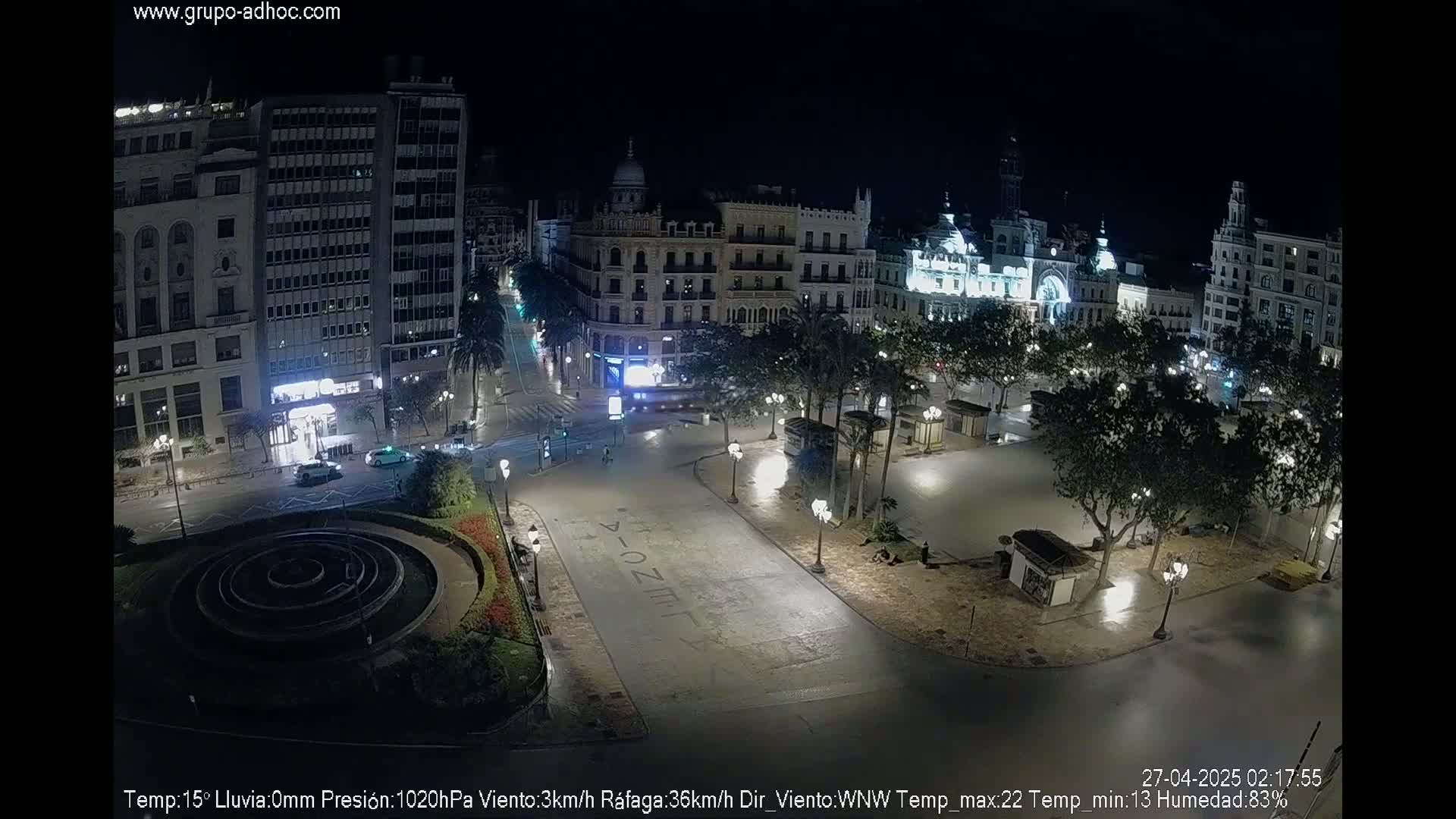 A nighttime view of a mostly empty city square surrounded by illuminated buildings, with a circular fountain in the foreground.