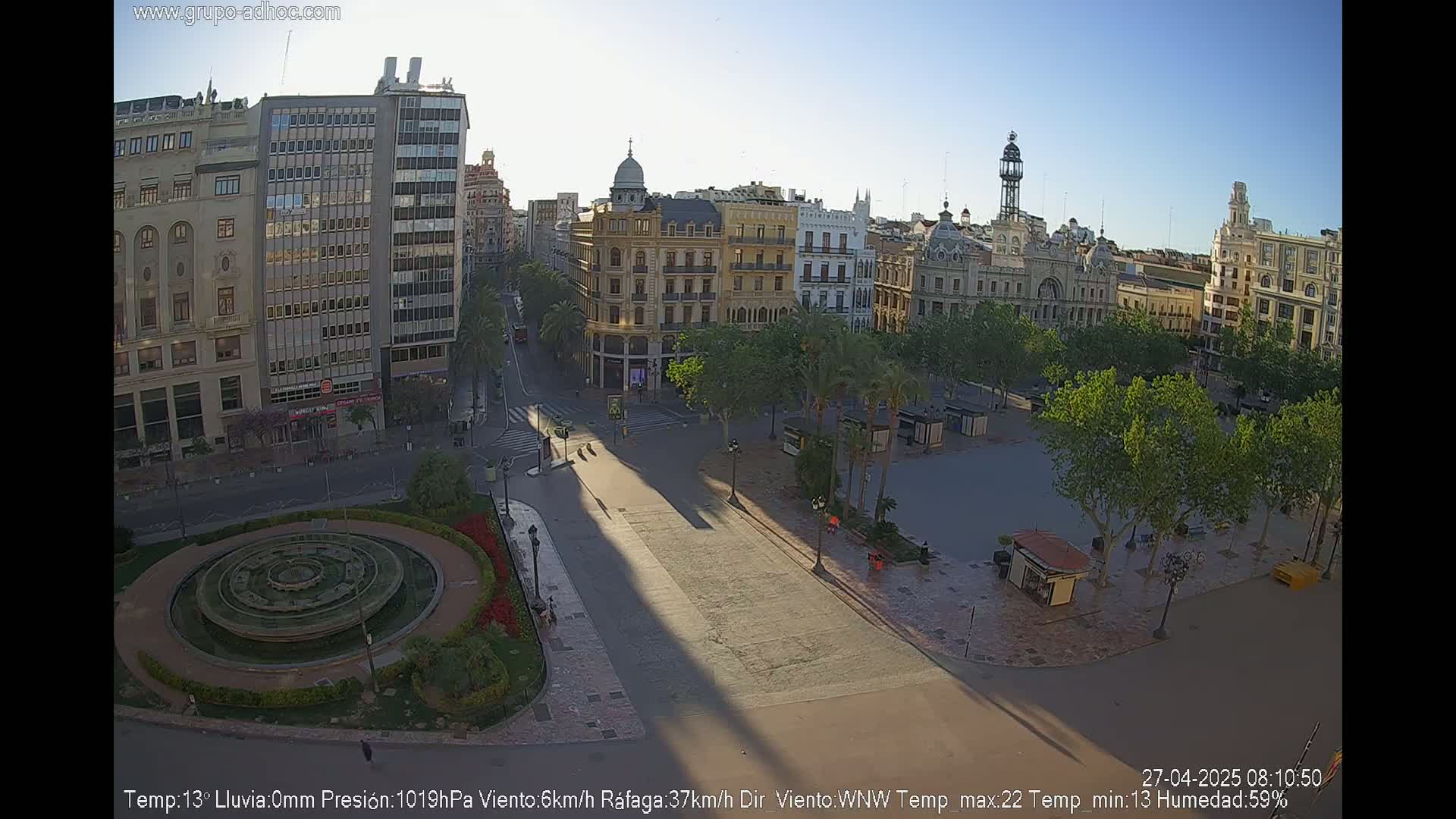 A mostly empty city square with a circular fountain and surrounding buildings is seen on a sunny morning.