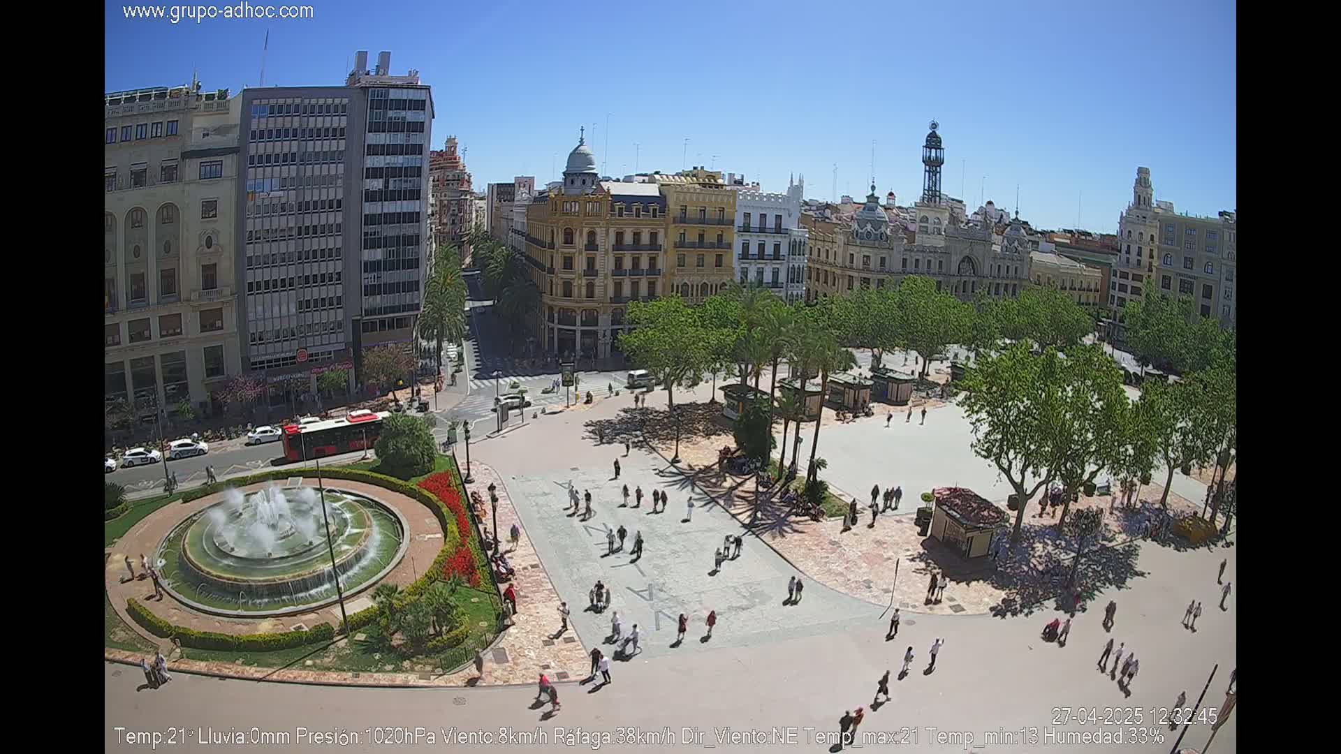 A sunny city square with a central fountain, surrounded by buildings and palm trees, shows numerous people walking around.