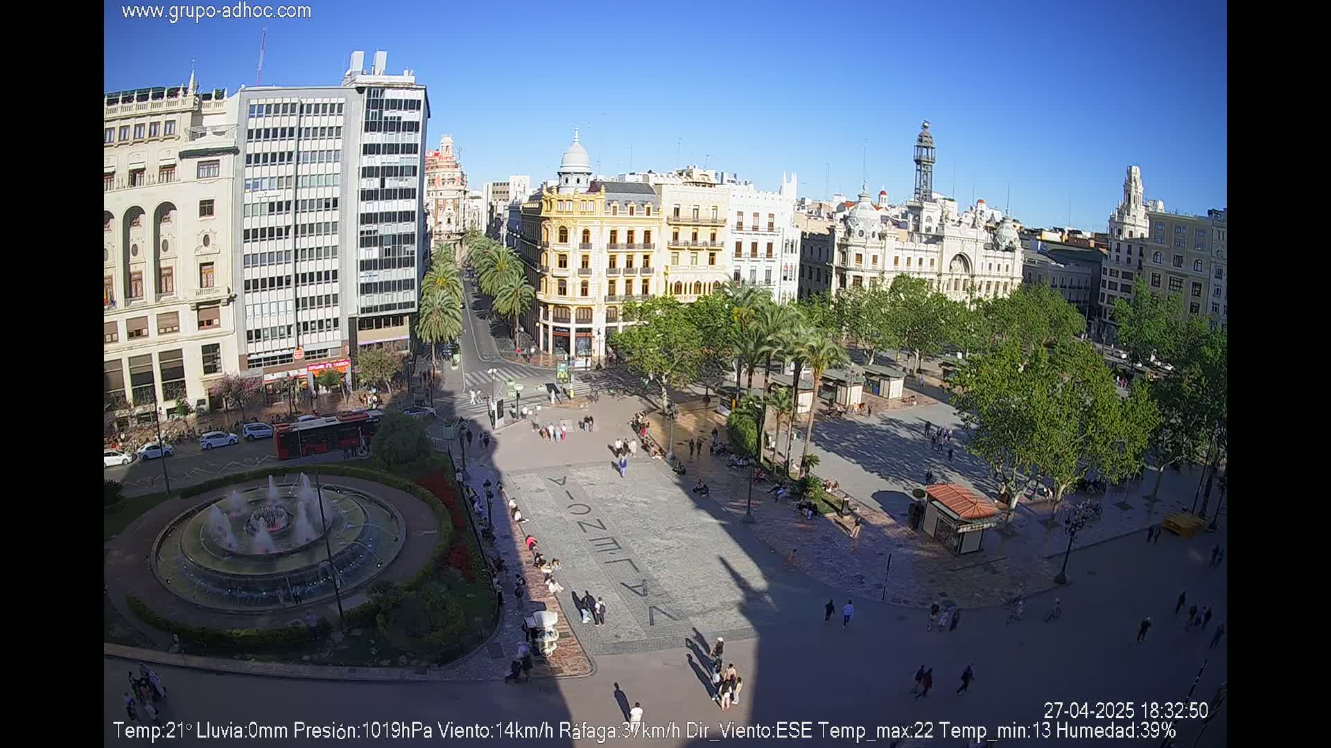 A sunny city square with a central fountain, surrounded by buildings and palm trees, and many people walking around.