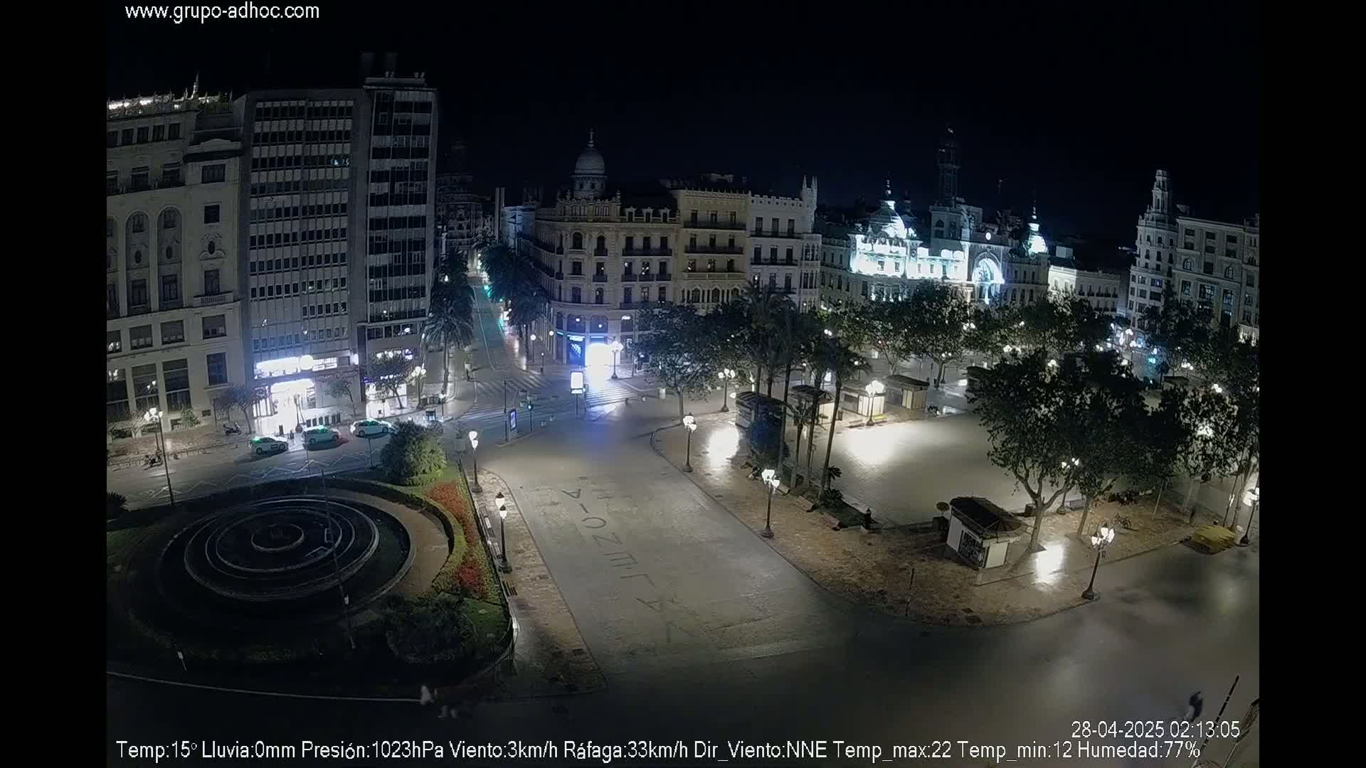 A nighttime, lightly raining view of a city square with several buildings and palm trees, illuminated by streetlights.