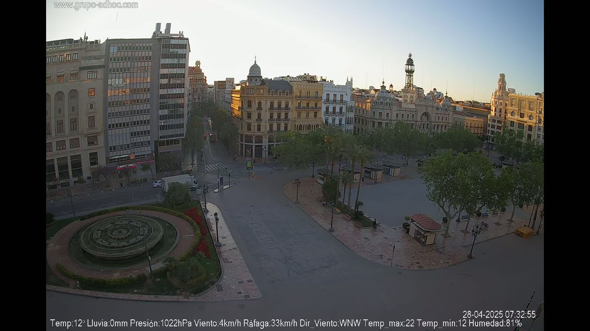 A mostly empty city square with a circular fountain, surrounded by several buildings under a clear, dawn sky.