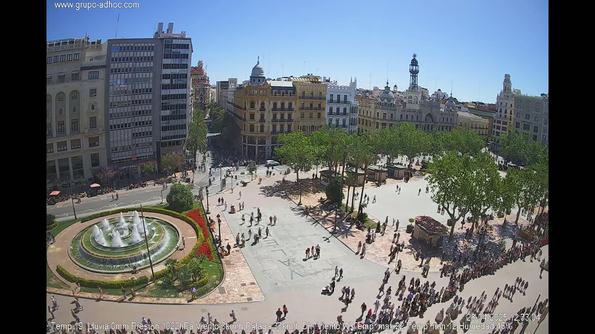 A sunny city square with a central fountain, surrounded by buildings and people, under a clear blue sky.