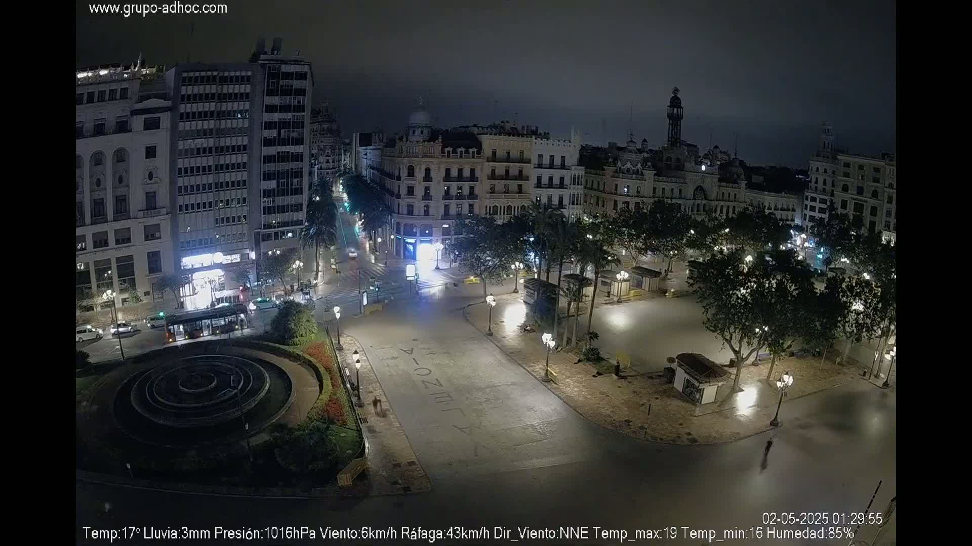 A nighttime, rain-slicked city square is sparsely populated with streetlights illuminating surrounding buildings and a circular fountain.