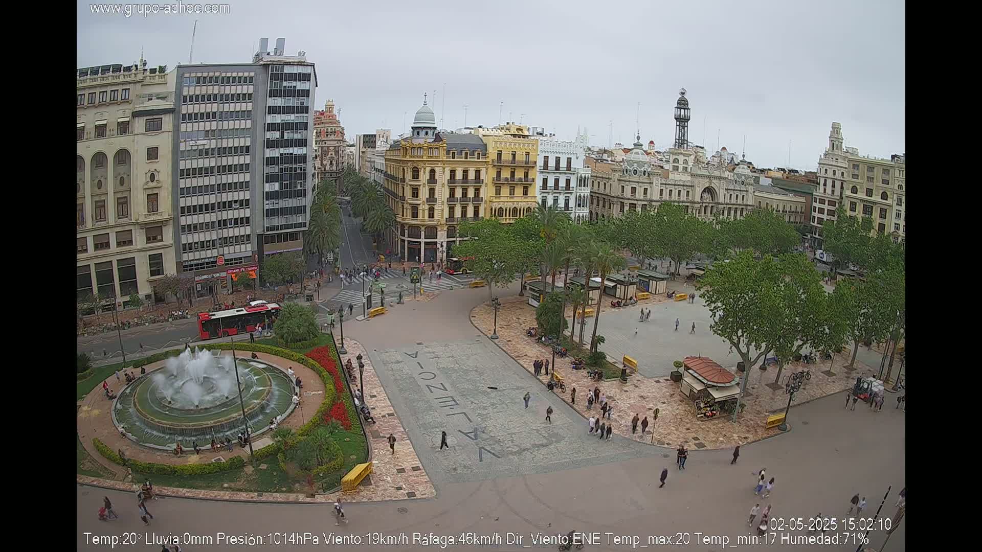 A city square with a fountain, surrounded by buildings and palm trees, on an overcast day.