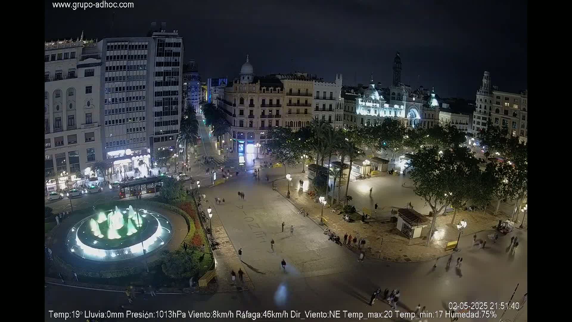 A nighttime aerial view of a city square with a lit fountain, surrounded by buildings and palm trees under a clear sky.