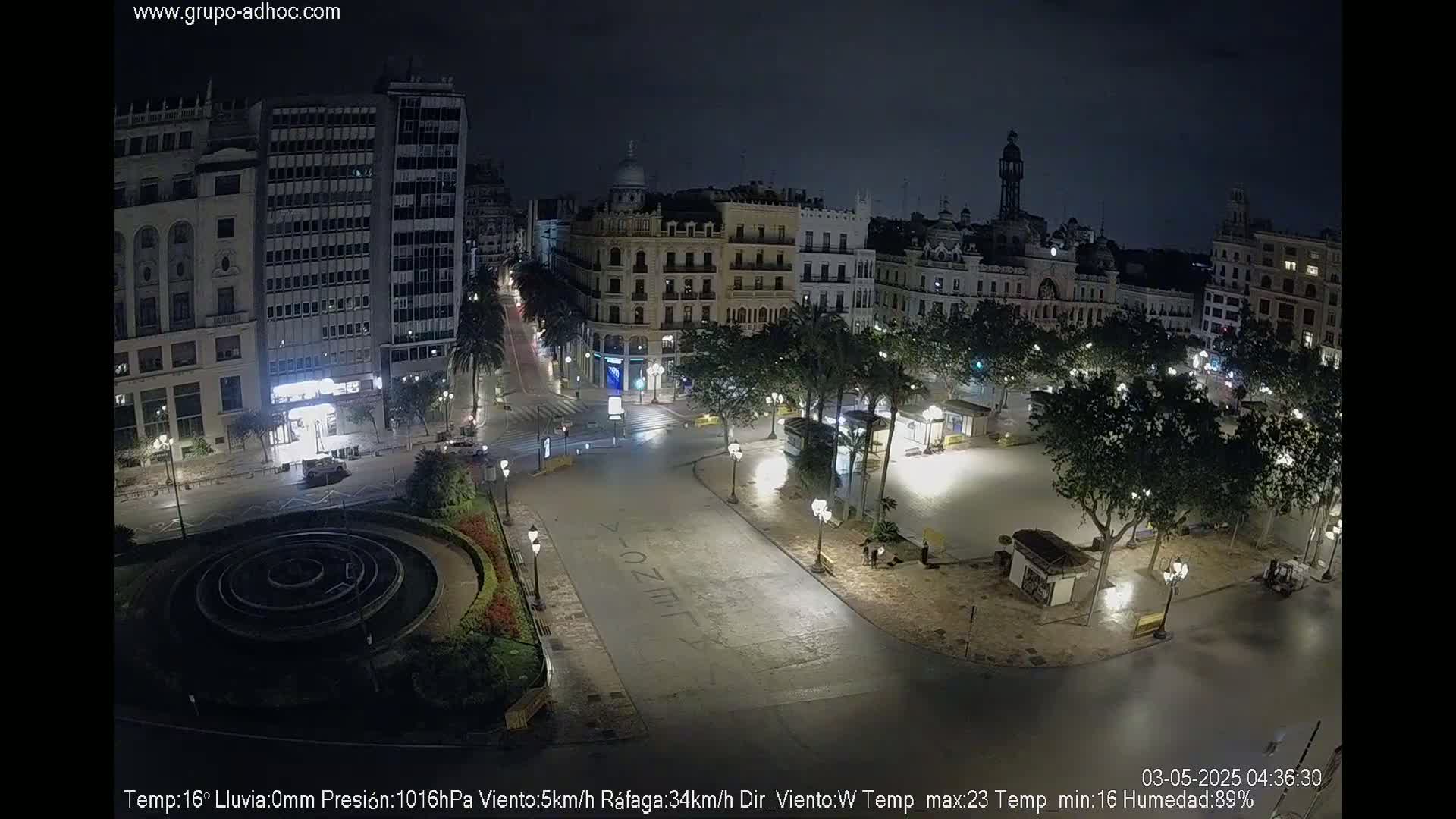 A nighttime, deserted city square with surrounding buildings, trees, and a circular fountain, under a clear sky.