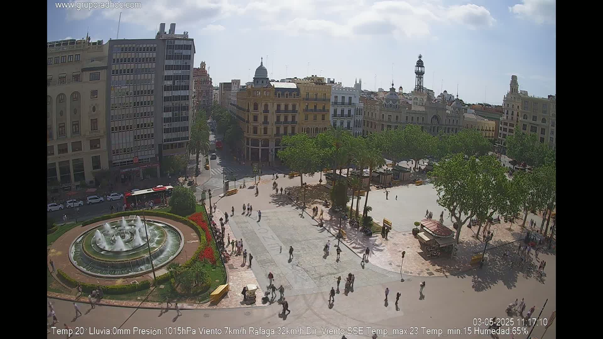 A sunny city square with a fountain, surrounded by buildings and trees, and people walking around.