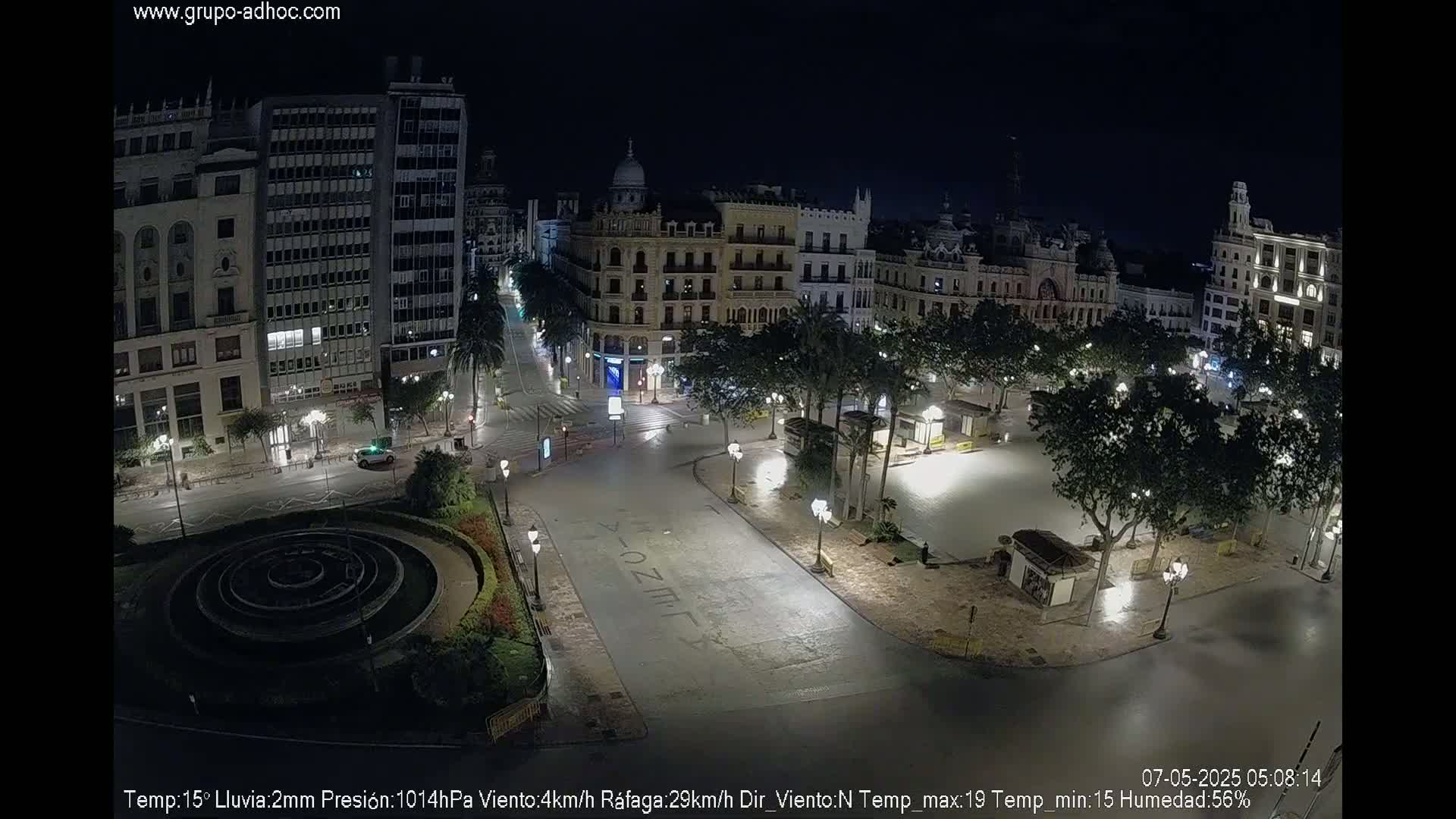 A mostly empty plaza and surrounding city streets at night, lightly raining.
