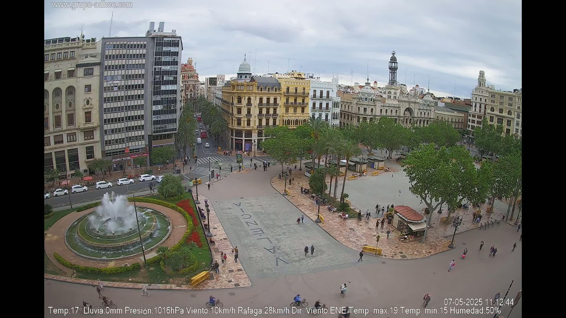 A city square with a fountain and several buildings is viewed from above on a cloudy day.