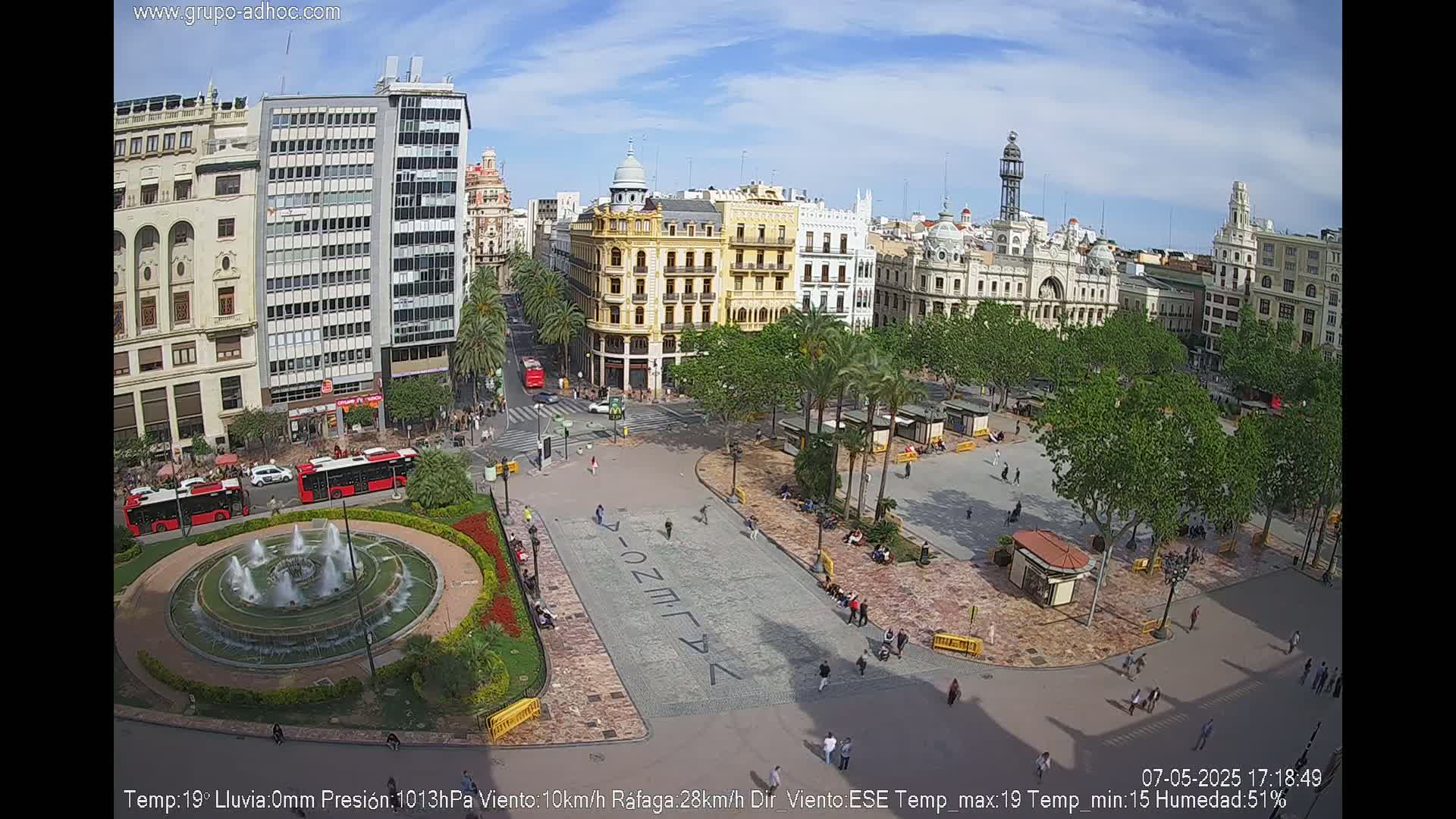 A city square with a large fountain, surrounded by buildings and palm trees, under a mostly sunny sky.
