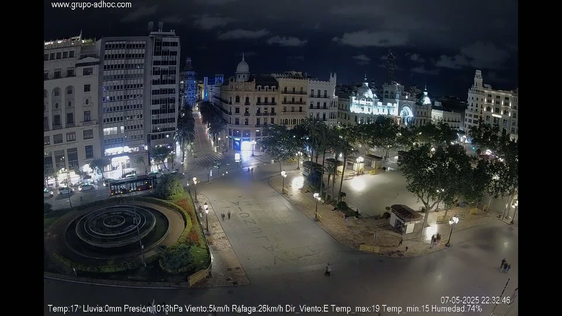 A nighttime, mostly clear view of a city square with buildings illuminated, a fountain, and sparse pedestrian traffic.